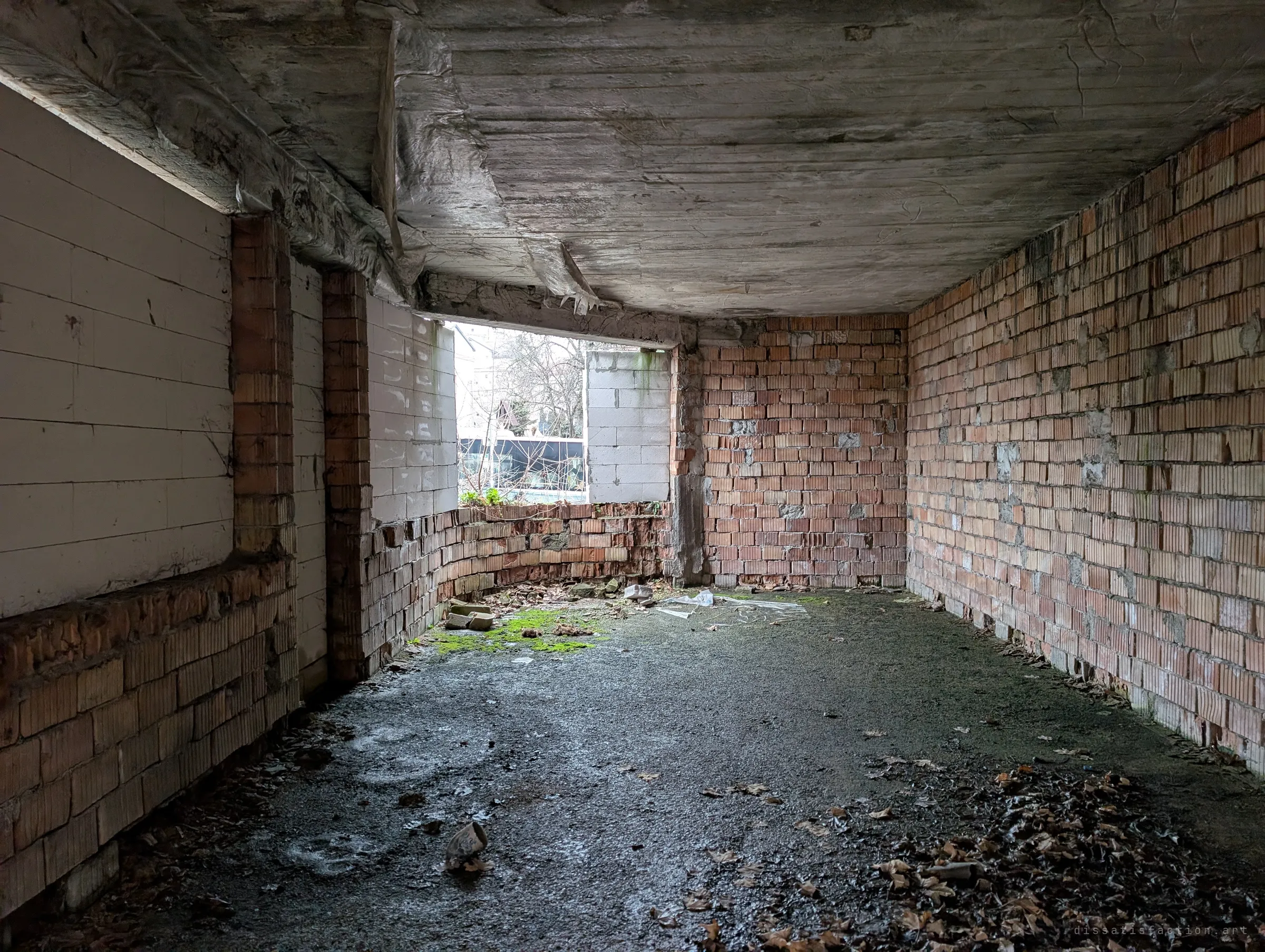 A low brick-walled room under a rough concrete ceiling, one window opening onto the street where a bus passes. Leaves and rubble drift across the wet floor.