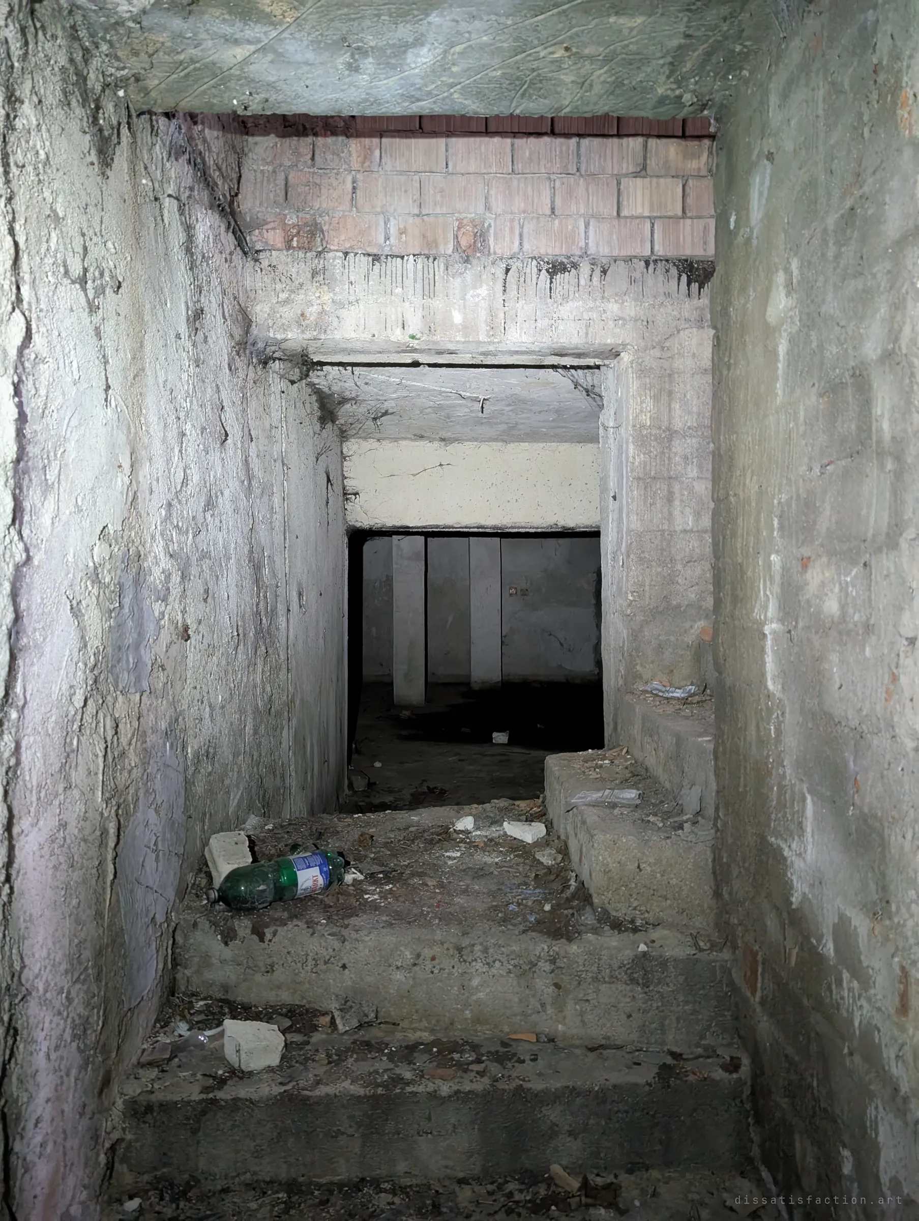 Concrete steps lead down into a torchlit basement opening, a discarded plastic bottle on the landing. Pillars wait in the dark room below.