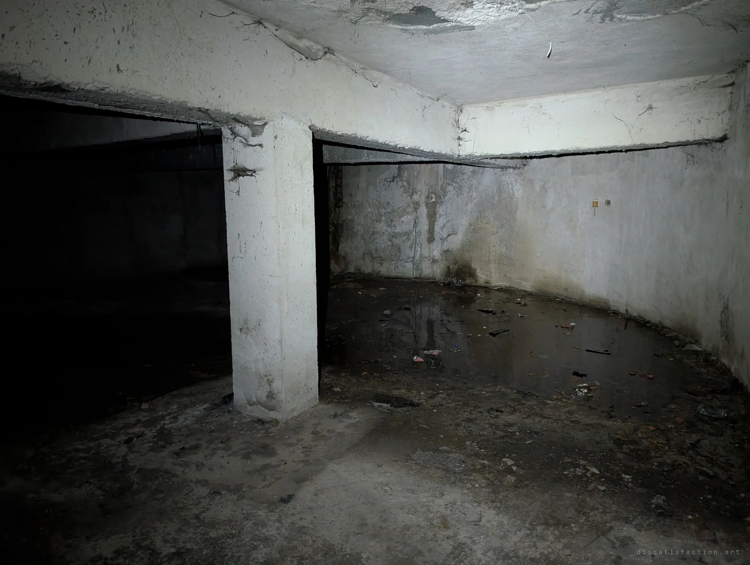 A single pillar in a pitch-dark basement, a curved wall wrapping around standing water littered with debris. The round footprint of the building continues underground.