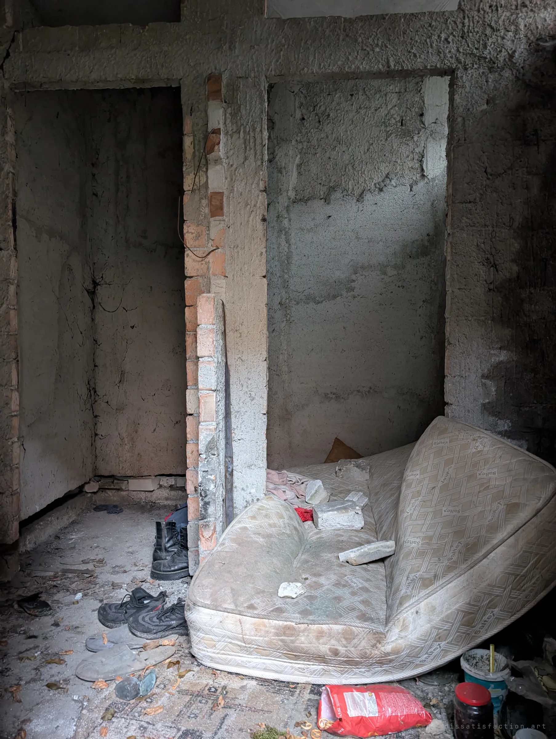 A folded, stained mattress, boots and shoes left in a rough concrete room. Signs that someone has been sleeping here.