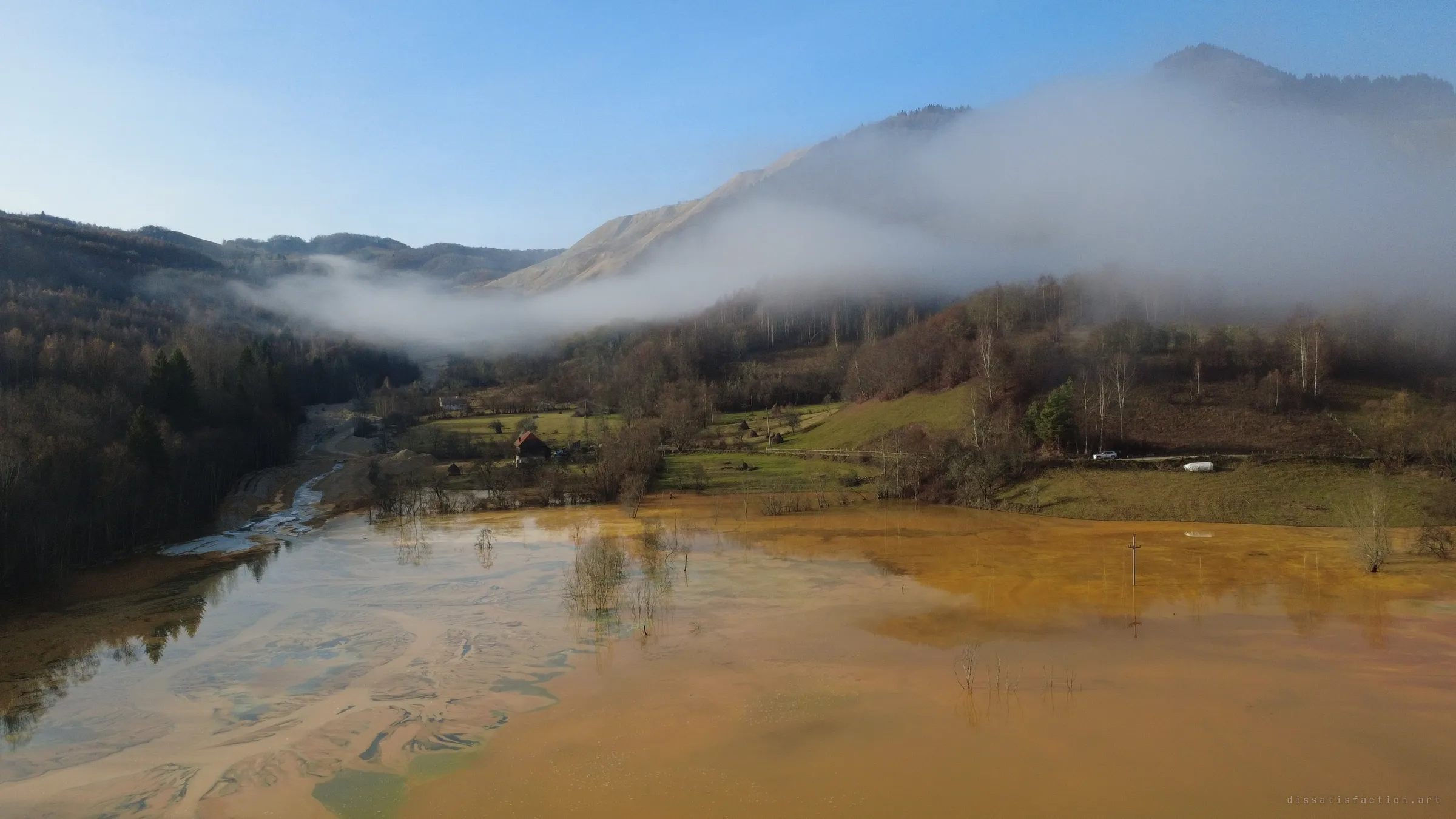 From above, orange tailings water floods a green valley floor up to a lone farmhouse, while fog hangs over bare, grey mine-waste slopes behind.
