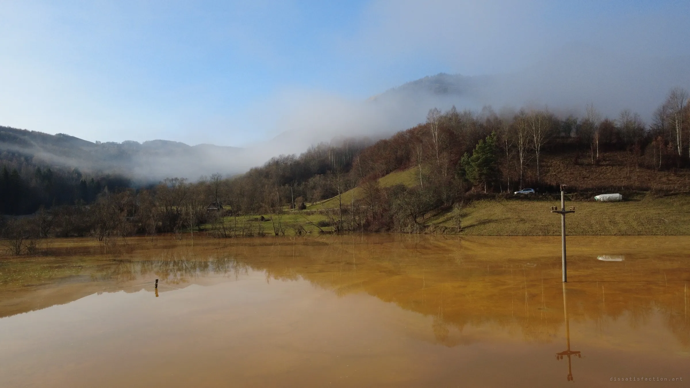 A power pole stands knee-deep in ochre water that has swallowed the valley floor, a parked car and a wrapped bale on the dry slope beyond, fog drifting across the hills.