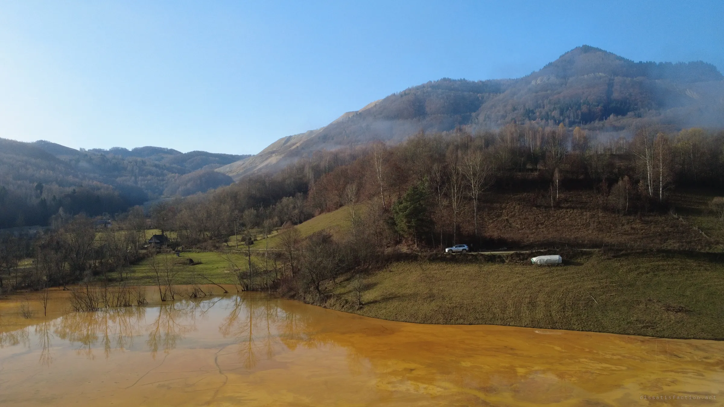 Clear morning light over a valley where orange water creeps up a grassy slope, bare trees standing in it and a pale mine-waste slope scarring the mountain behind.