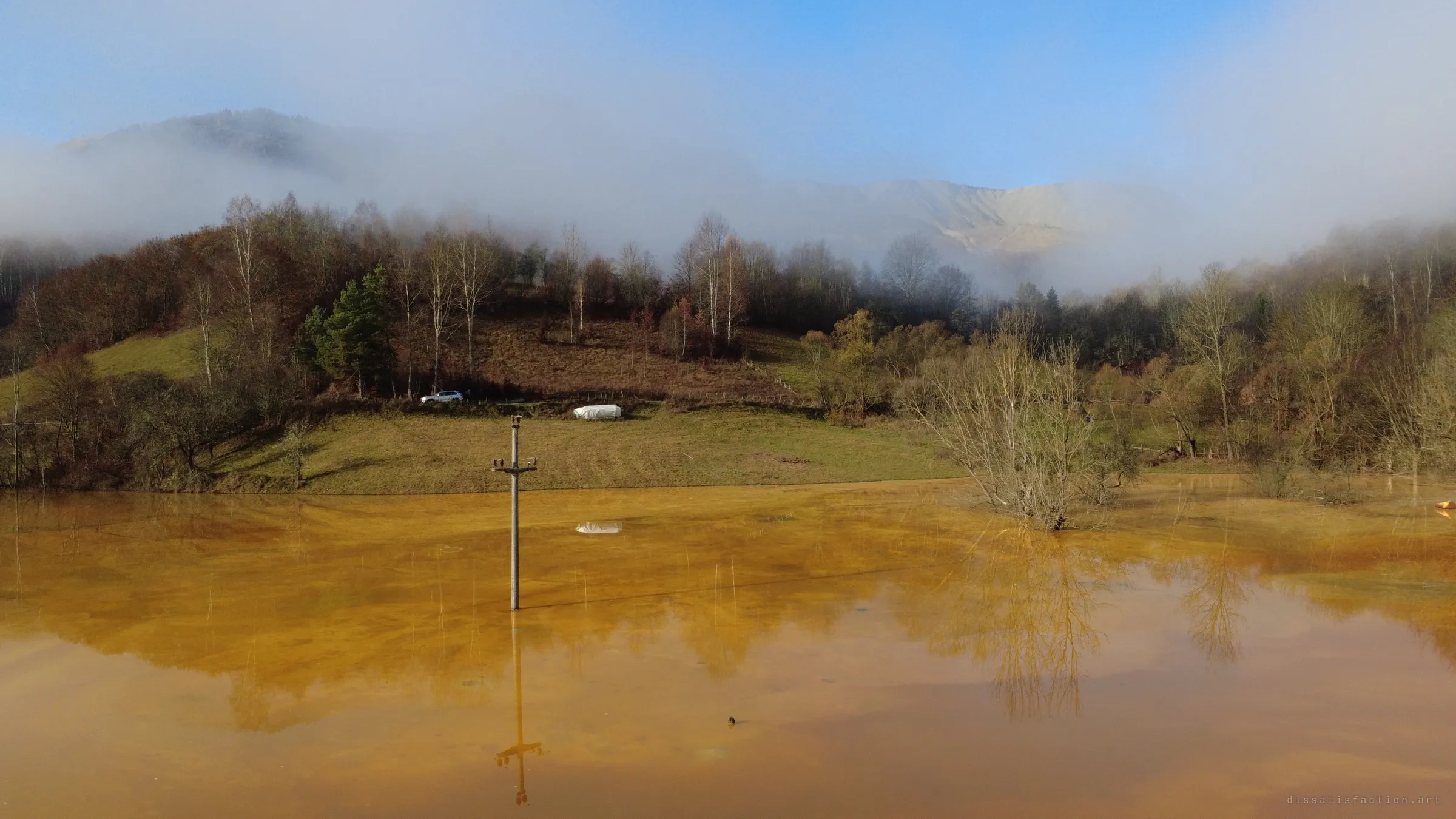 A half-sunk power pole and a drowned tree stand in mustard-orange water, a grey waste slope showing through the fog on the ridge behind.