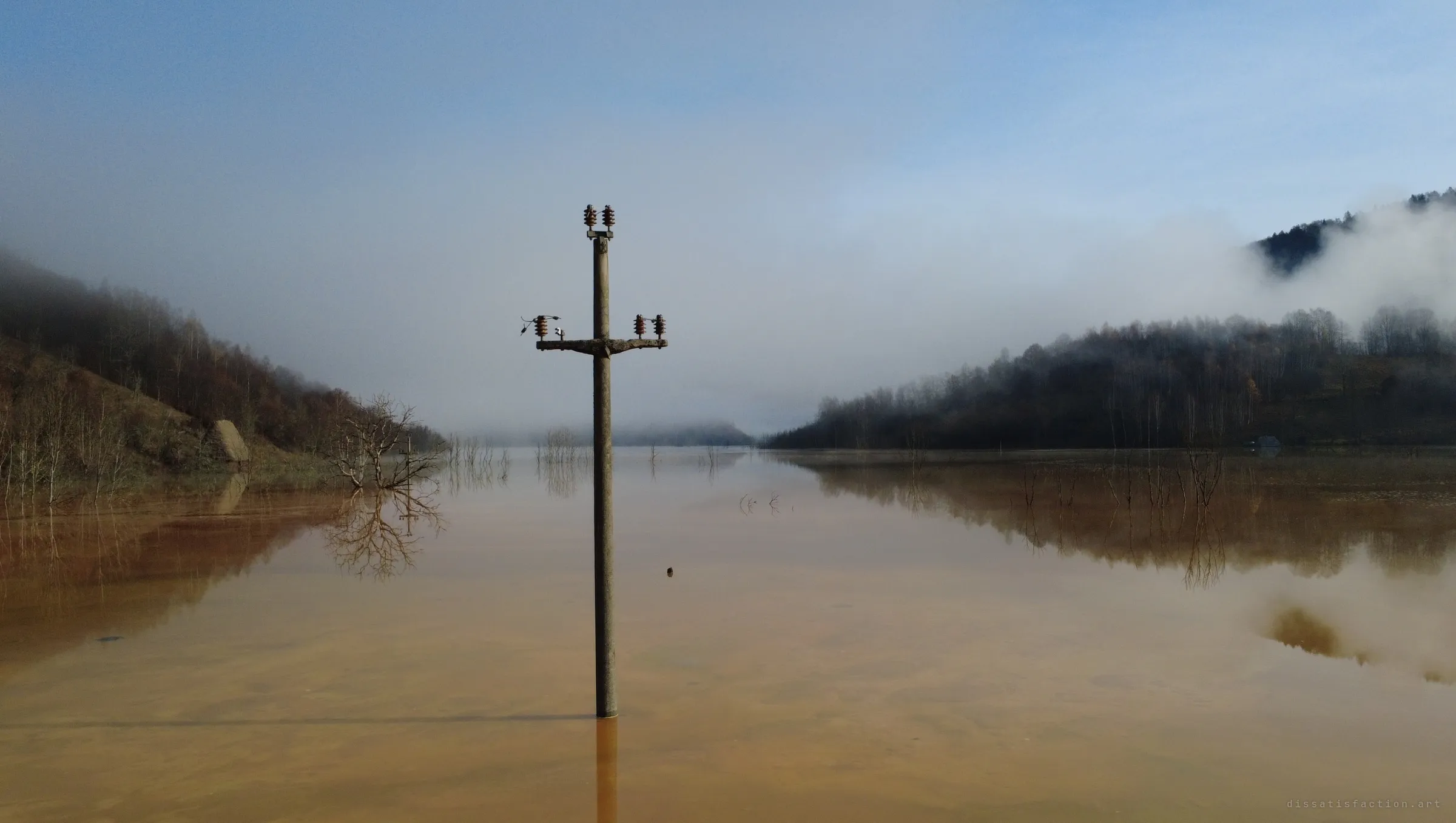 A lone power pole, insulators still in place, rises from a flat rust-coloured lake that fills the valley and fades into fog.