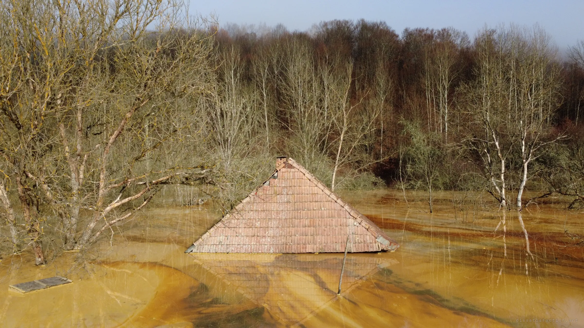 Only the peak of a tiled roof and its chimney stay above the ochre water, a leaning post and floating boards beside it, bare birches behind.