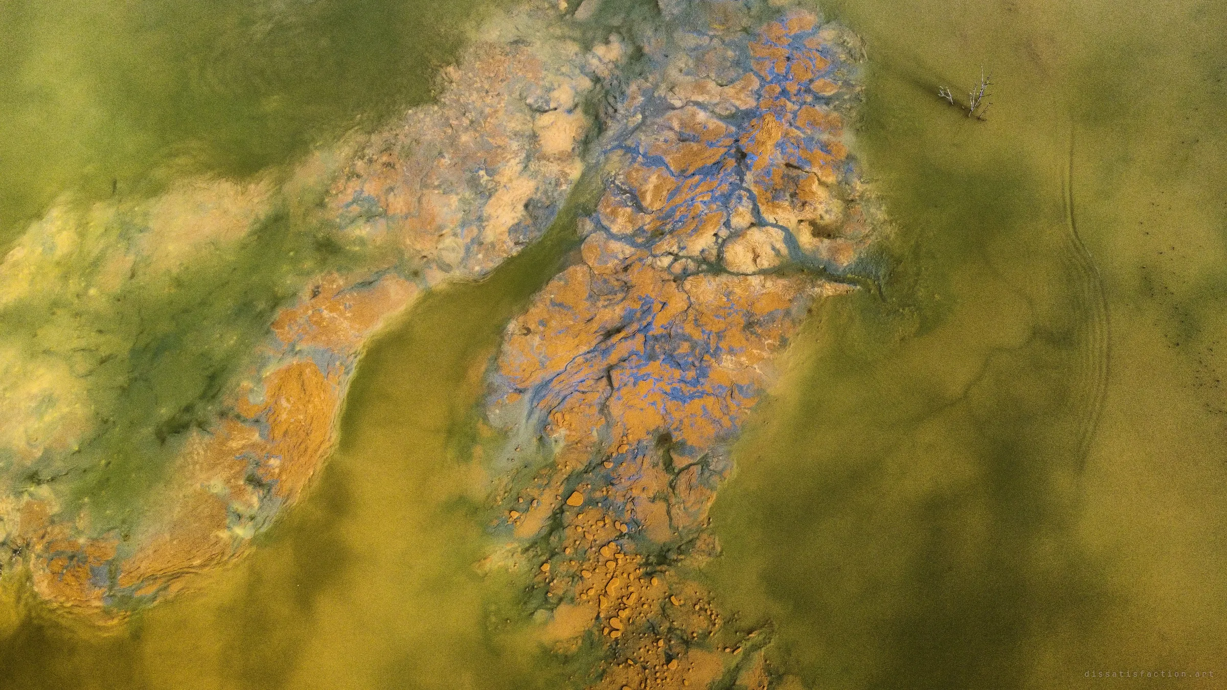 Looking straight down on crusted orange and violet mineral banks threading through murky green-yellow water.