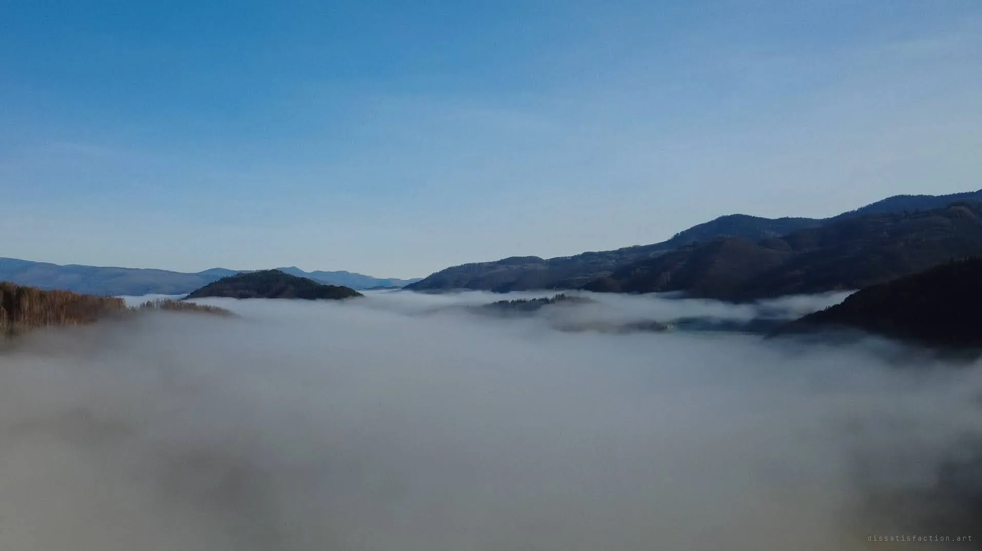 Aerial shot above a sea of fog filling a mountain valley, only forested ridges breaking through under a clear blue sky. The valley that hides the site stays buried under cloud.