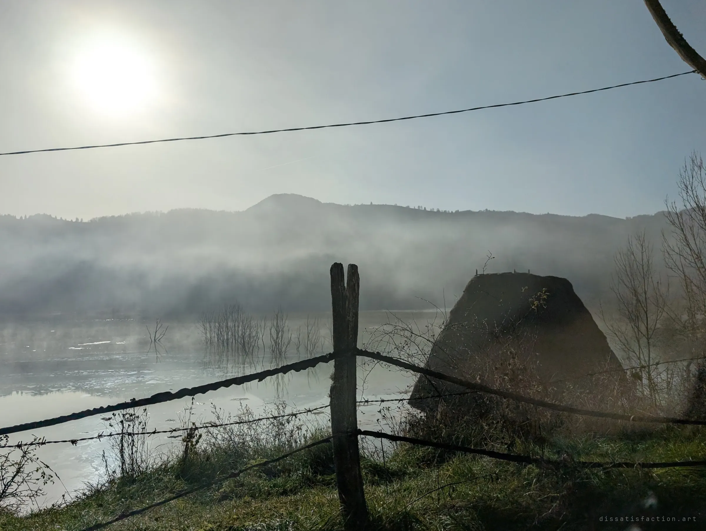 A barbed-wire fence and a haystack on the shore of a misty lake at sunrise, dead trees poking out of the water. Morning fog hangs over the flooded valley and the hills behind it.