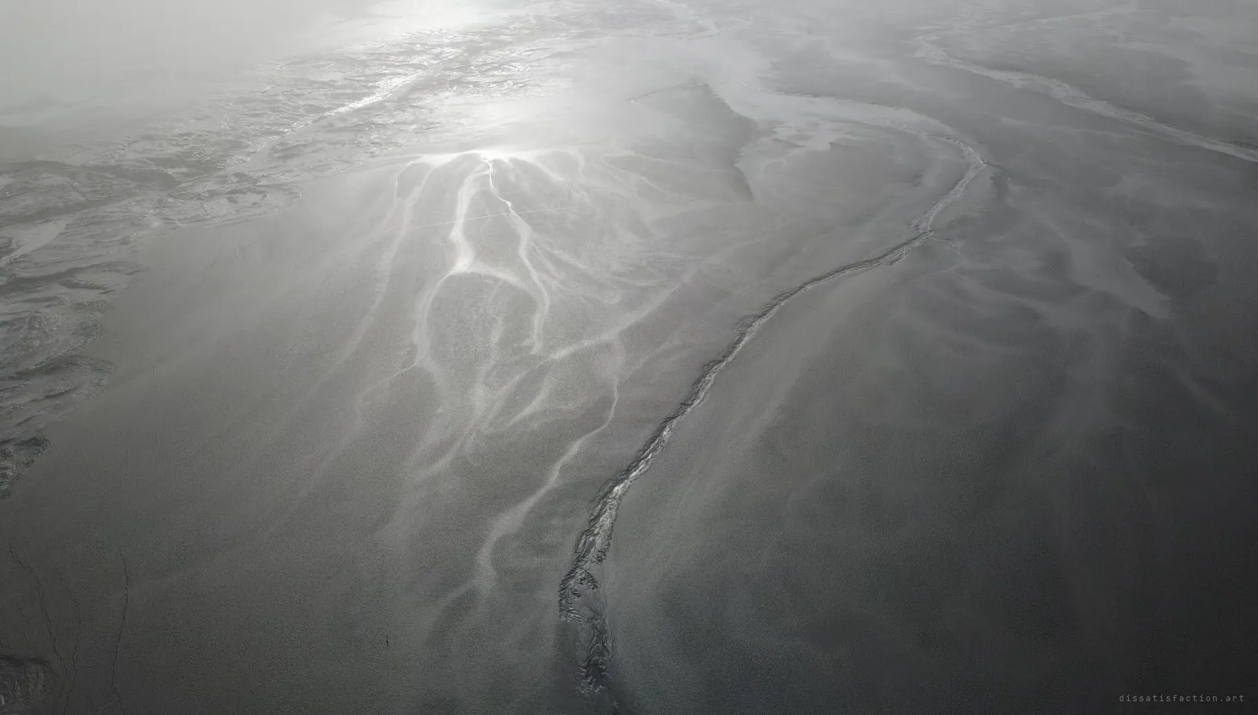 Looking down on a plain of grey mining sludge, pale channels branching across it like veins toward a glint of light. The waste has swallowed everything that used to be here.