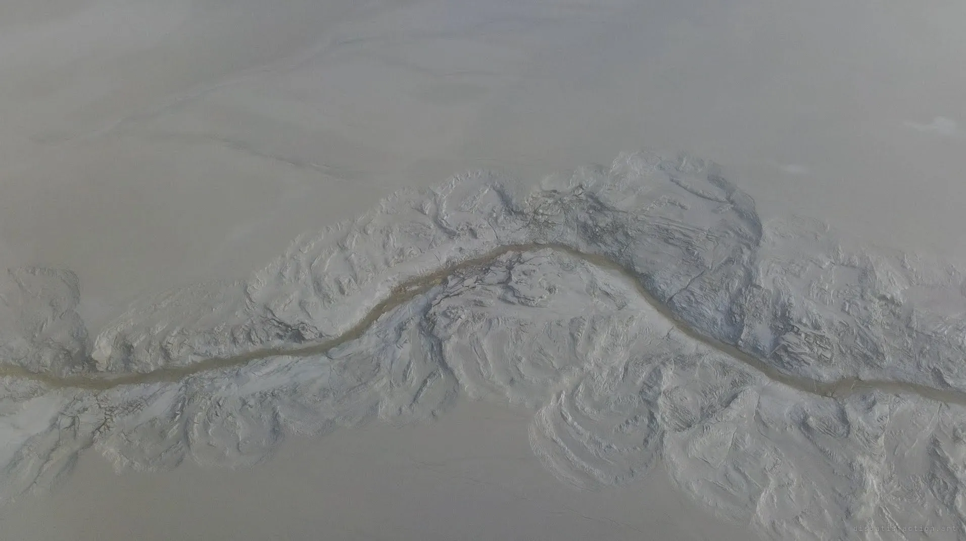 Top-down view of a thin brown stream winding through cracked, folded ridges of pale grey sludge. It reads almost like a mountain range seen from orbit.