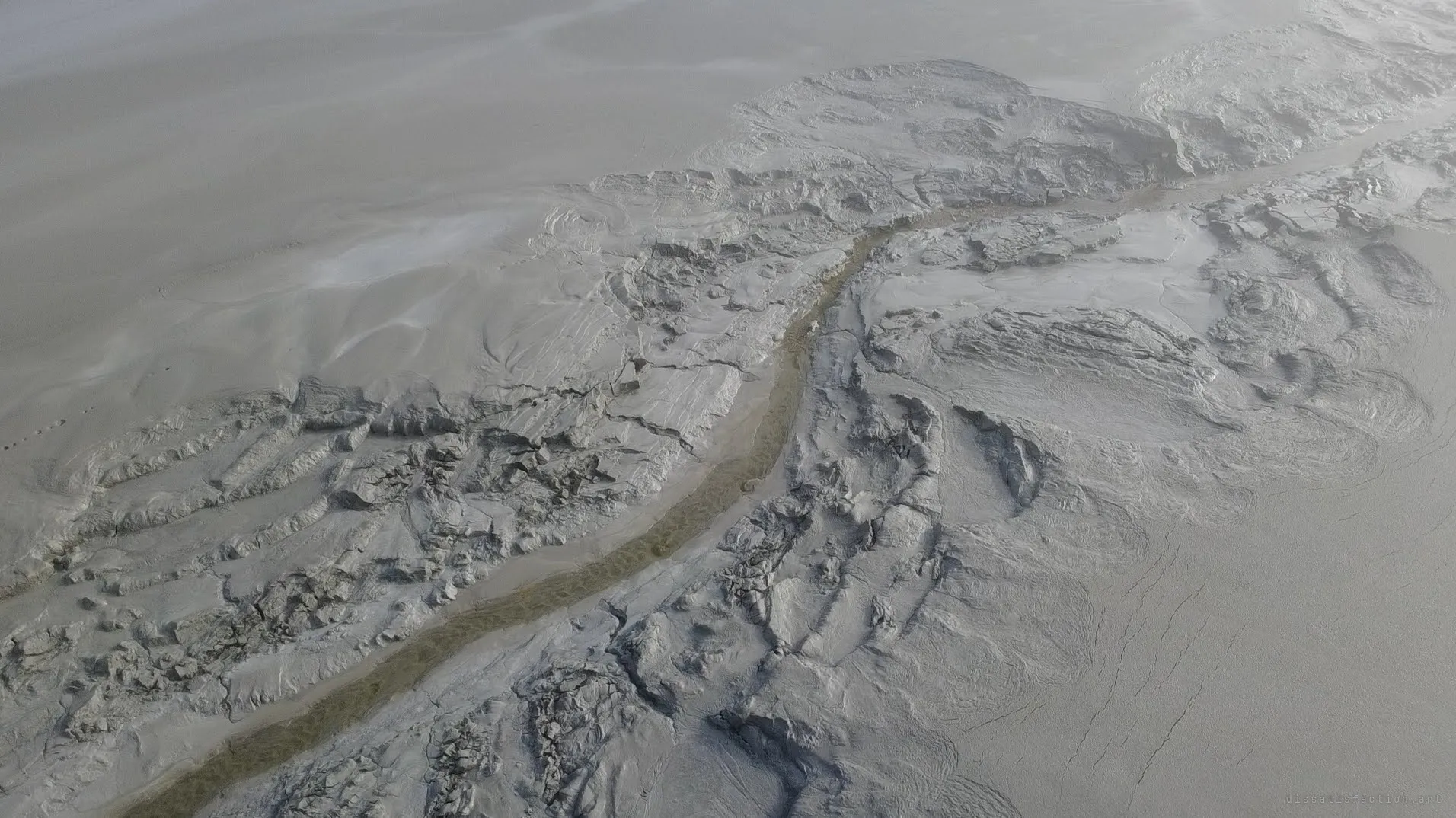 A murky green-brown channel cuts diagonally through slabs of buckled grey sludge. Up close, the mining waste looks like a frozen landscape of its own.