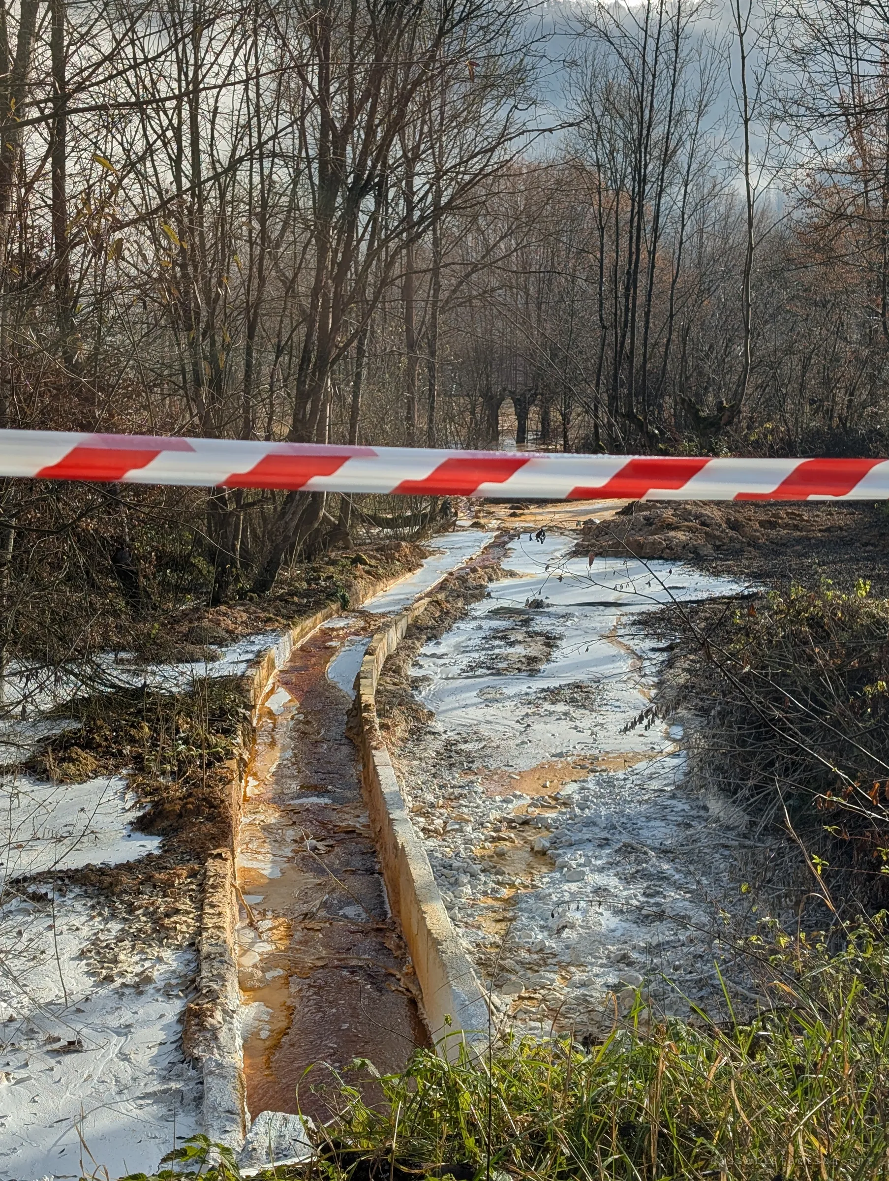 Red and white barrier tape stretched across a concrete runoff channel carrying rust-orange water, the ground on either side crusted white. The toxic outflow runs off into bare winter woods.