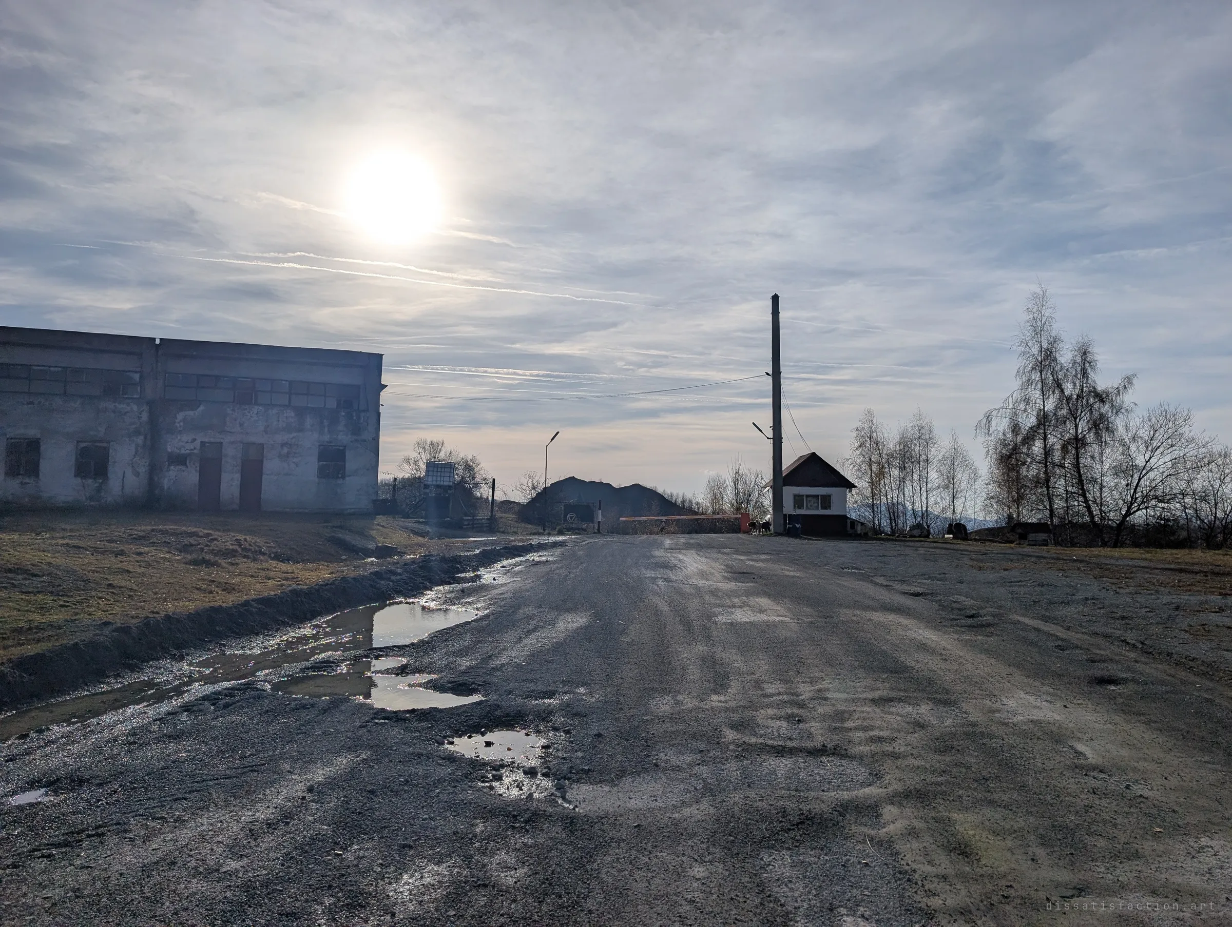 A potholed gravel road leads to a gatehouse with a lift barrier beside a weathered two-storey workshop, the low sun hazy above. It is the way in to the working mine next to the site.