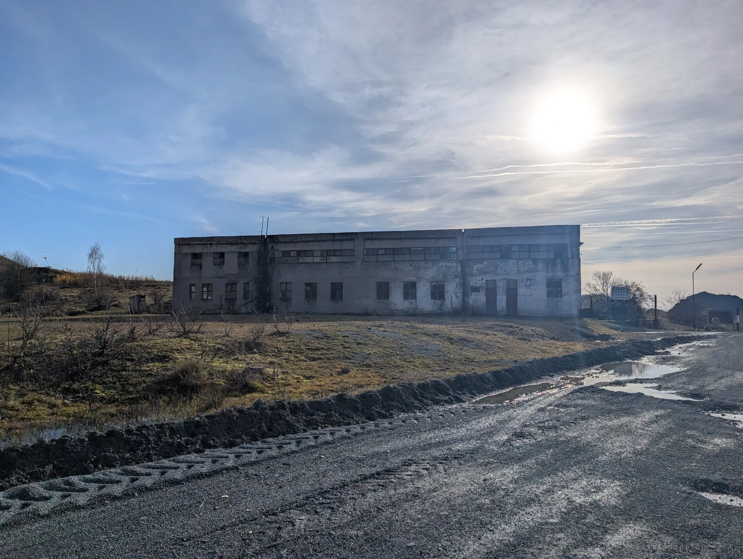 A long, weathered concrete workshop with rows of broken windows stands beside a muddy gravel road, backlit by a hazy sun. Tyre tracks and puddles show the road is still in use.