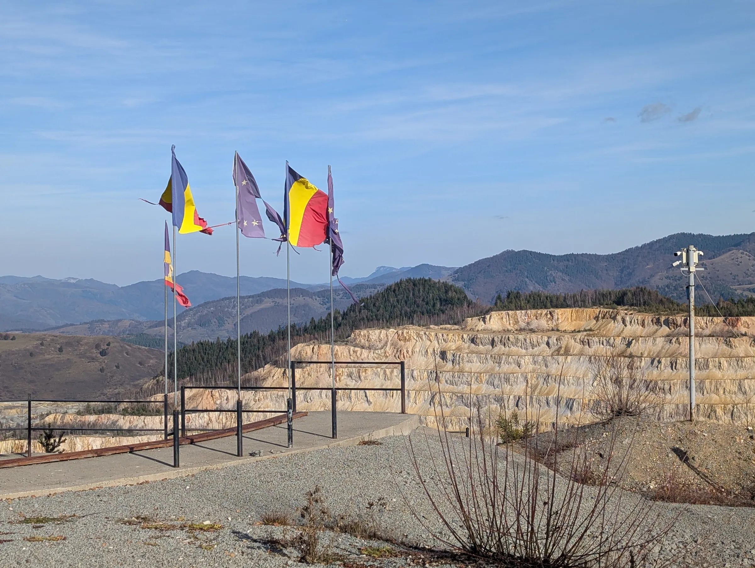 Tattered national and EU flags fly over a railed viewpoint at the edge of an open-pit mine, its pale stepped walls glowing in the sun. Forested mountains roll away behind.