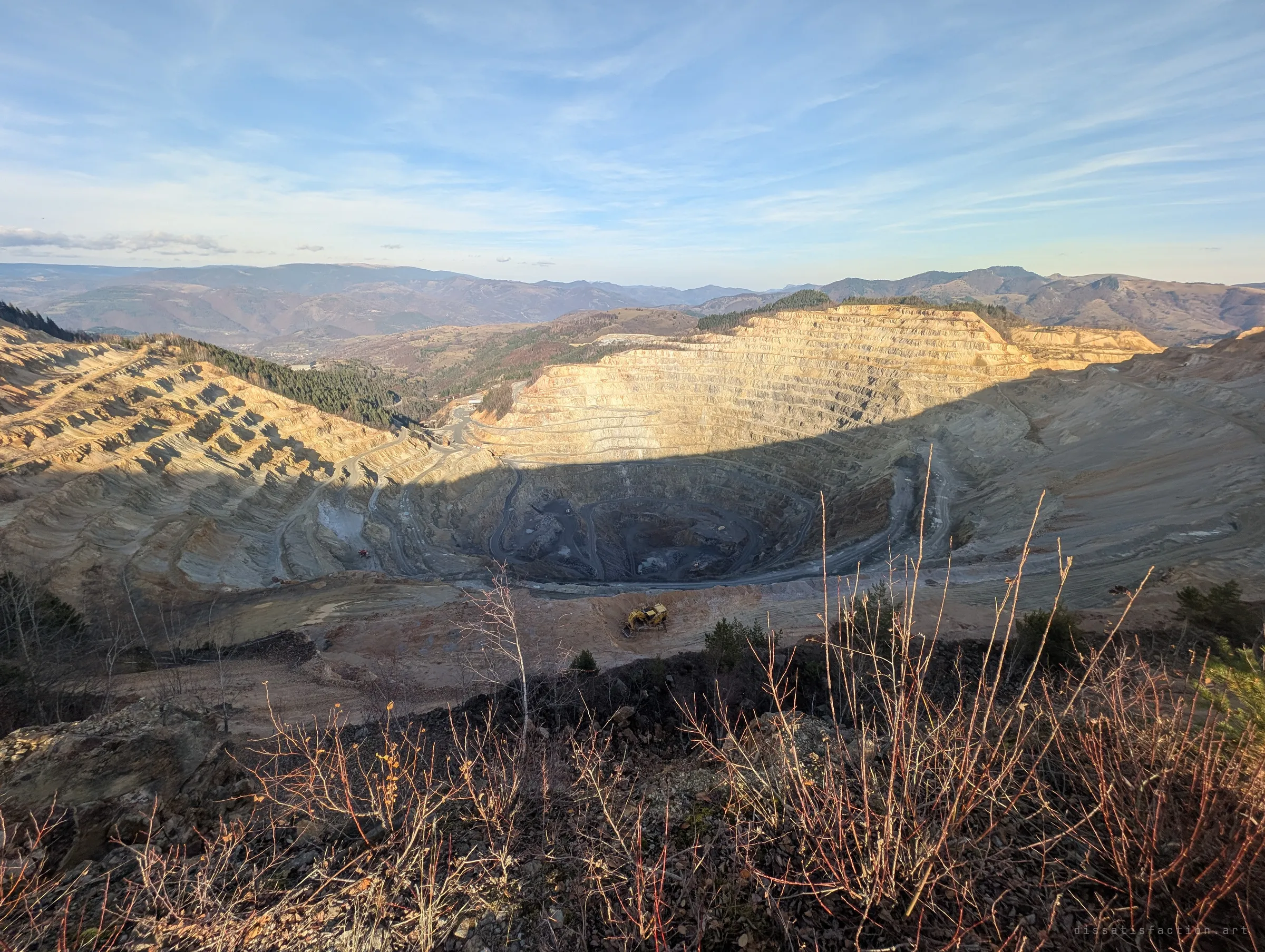 A vast open-pit mine spirals down into shadow, its terraced walls lit gold on the far side, with an excavator parked on the rim. The hills beyond fade into a hazy mountain horizon.