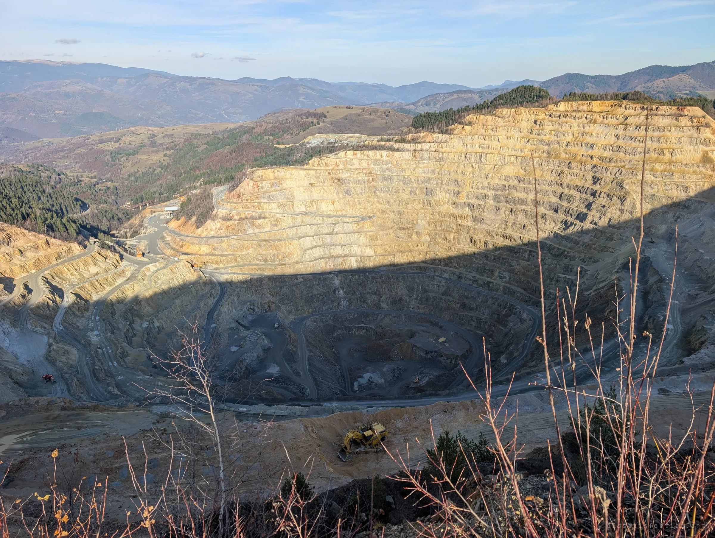 The stepped walls of the open pit catch the late sun while its floor sits in shadow, a yellow excavator working the near rim. The mine is still active, still producing the waste that buried the valley.