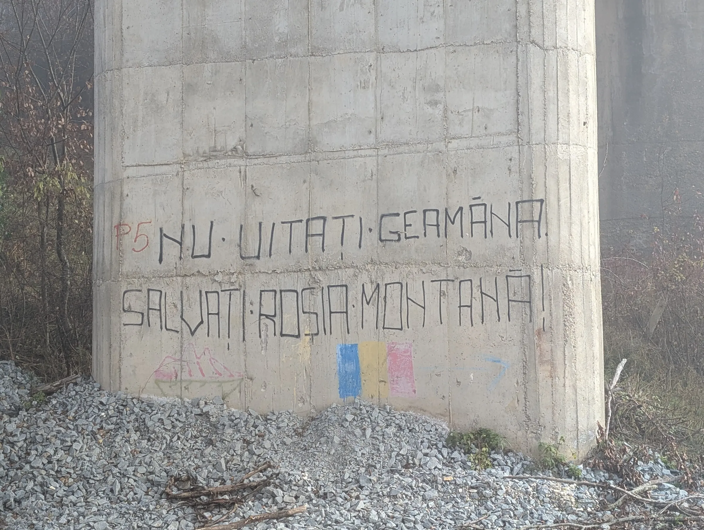 Hand-painted protest graffiti on a concrete bridge pillar, with a chalk tricolour and a drawing of mountains underneath. Someone wants the buried village remembered.