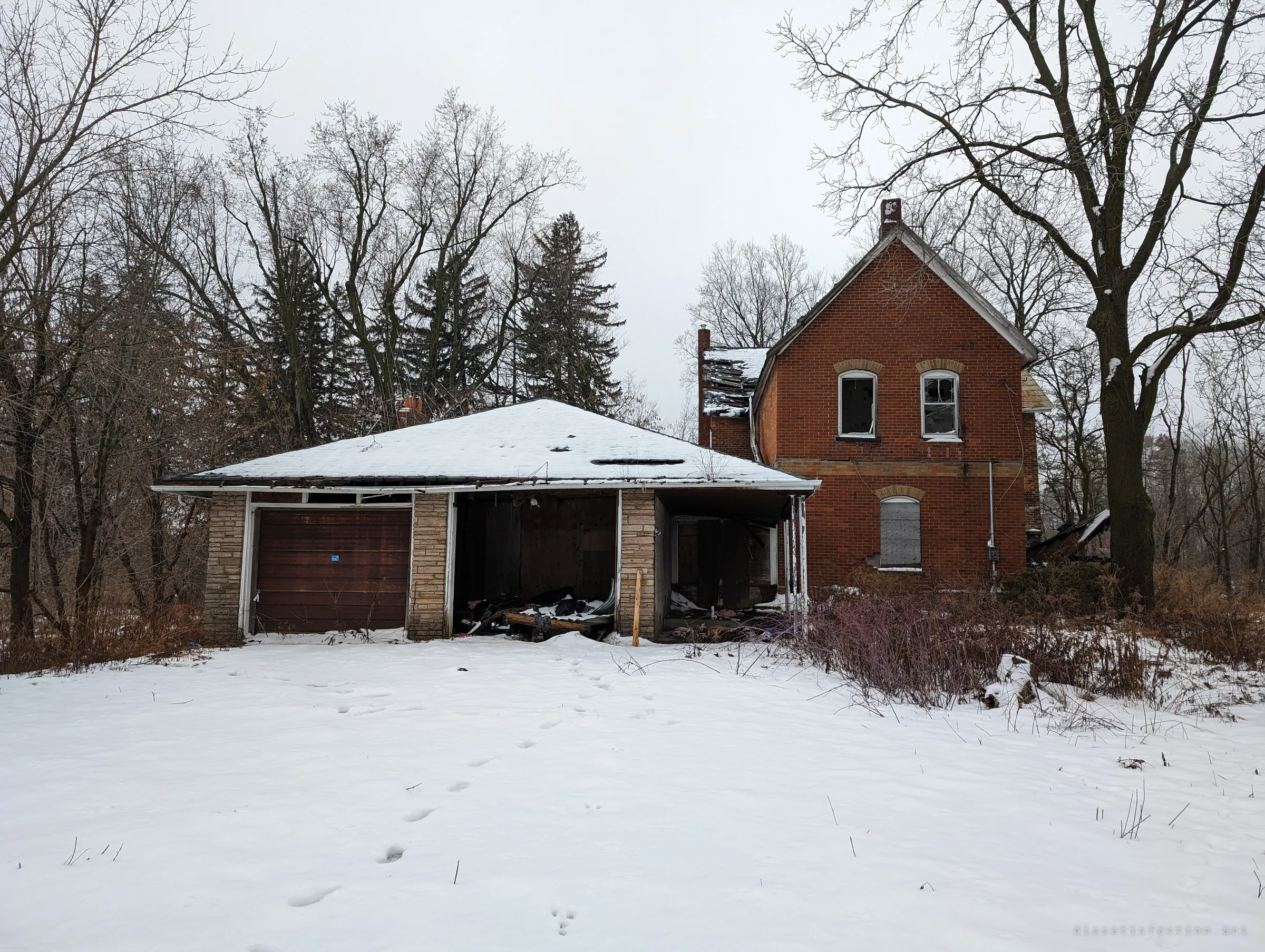 A two-storey red-brick farmhouse with a snow-covered, sagging garage in front, its bays open and full of debris. Footprints in fresh snow lead up to the empty building under a grey winter sky.