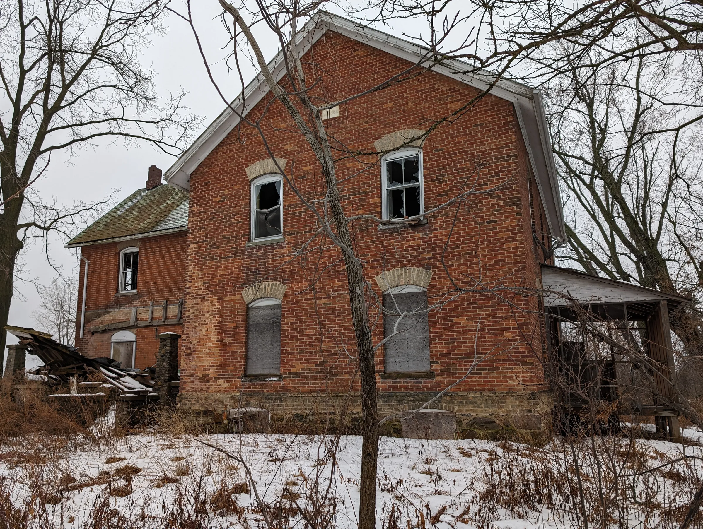 The tall brick gable end of the farmhouse, upper windows smashed and lower ones boarded, with a collapsed porch and a caved-in side structure. Bare saplings stand in the snowy weeds in front.