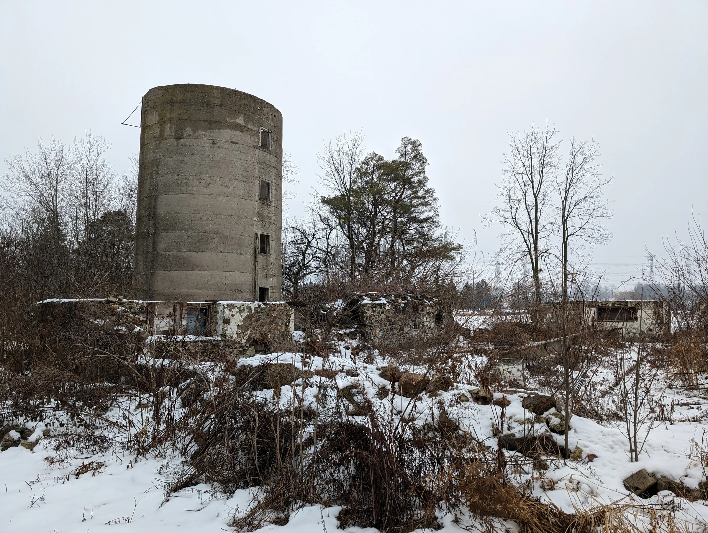 A weathered concrete silo rises behind the crumbling fieldstone foundations of a lost barn, half buried in snow and dead brush. The ruins show how much of the farm has already gone.