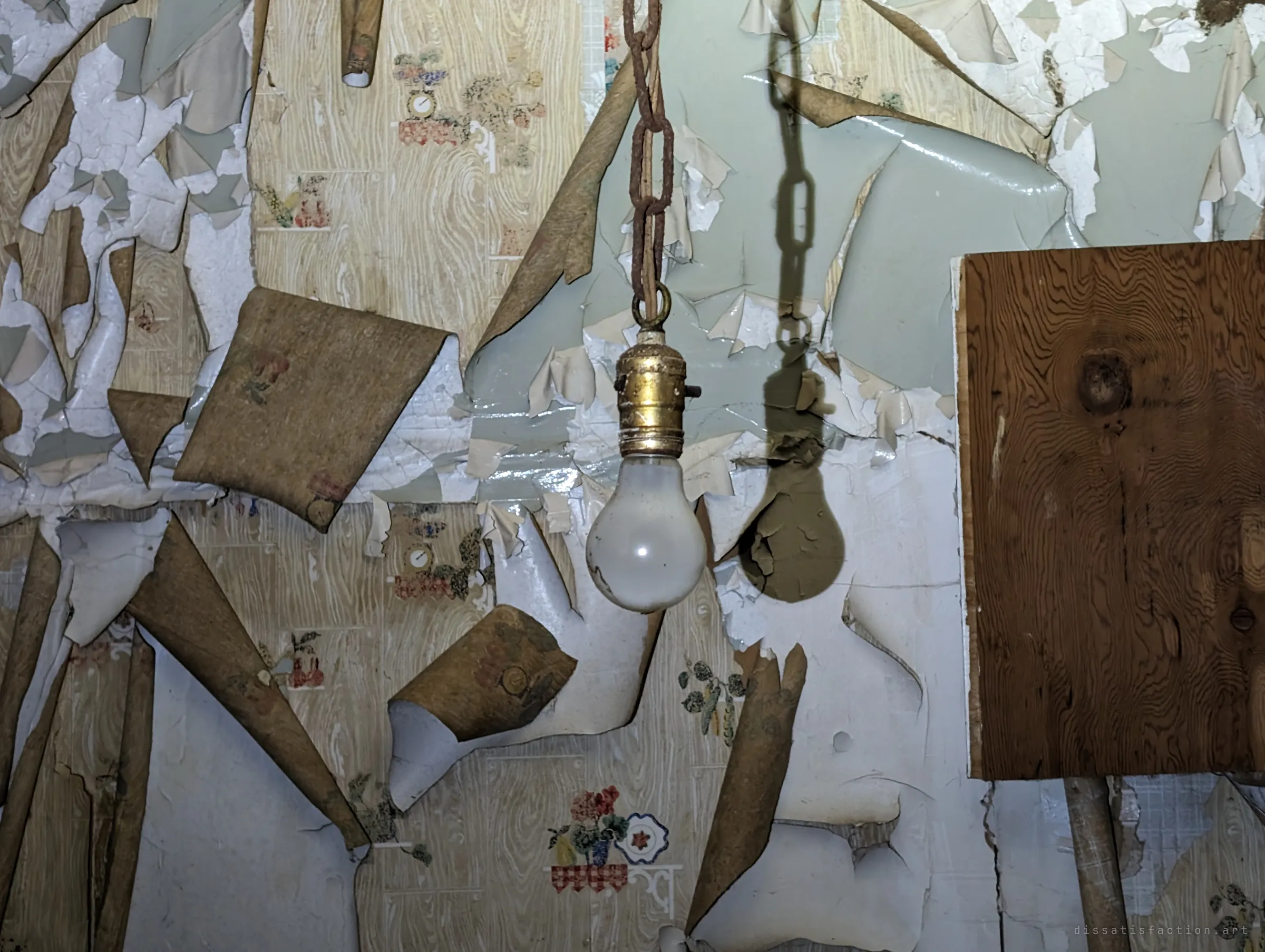 A bare bulb hangs from a rusted chain in front of a wall where layers of fruit-pattern wallpaper and pale green paint peel away in curling sheets. The bulb's shadow falls across the flaking wall beside a board.