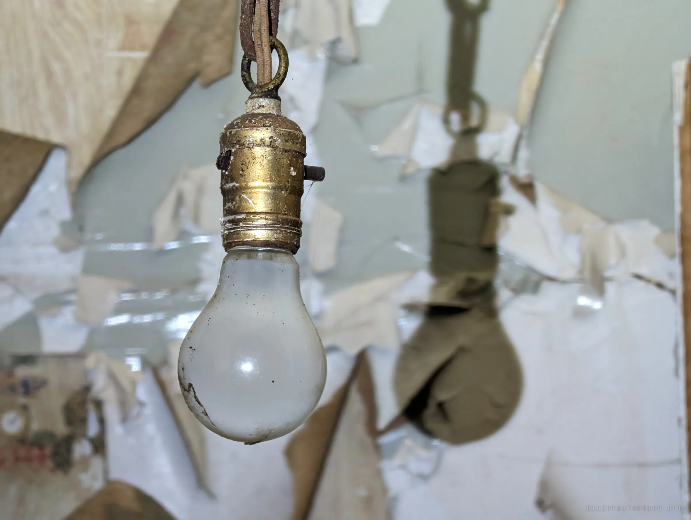 Close-up of the same frosted bulb in its tarnished brass socket, sharp against a softly blurred wall of peeling paint. Its long dark shadow echoes it on the right.