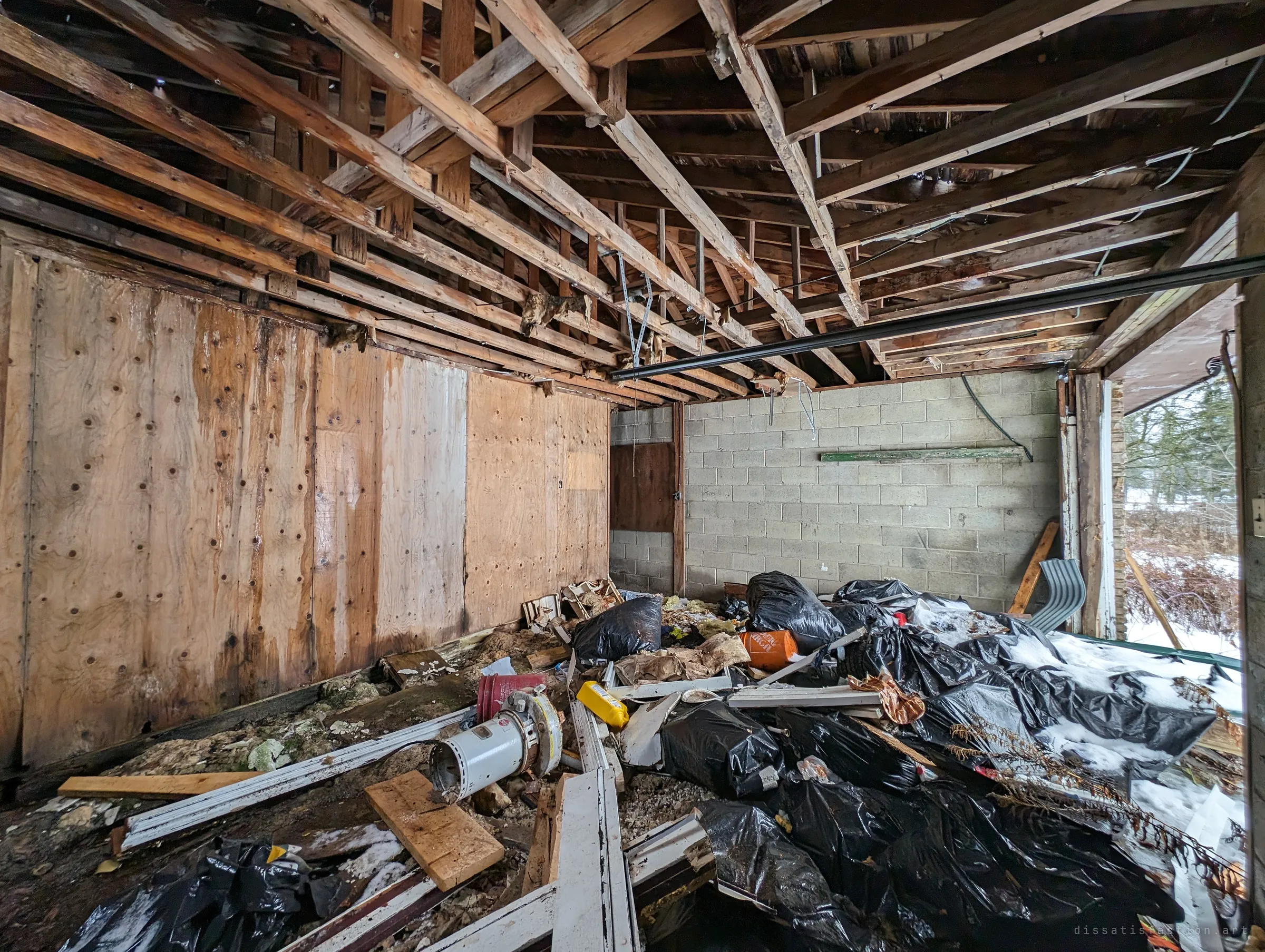 A gutted garage with bare roof joists, stained plywood walls and a heap of black bin bags and broken window frames. Snow drifts in through the open side, showing how exposed the building has become.