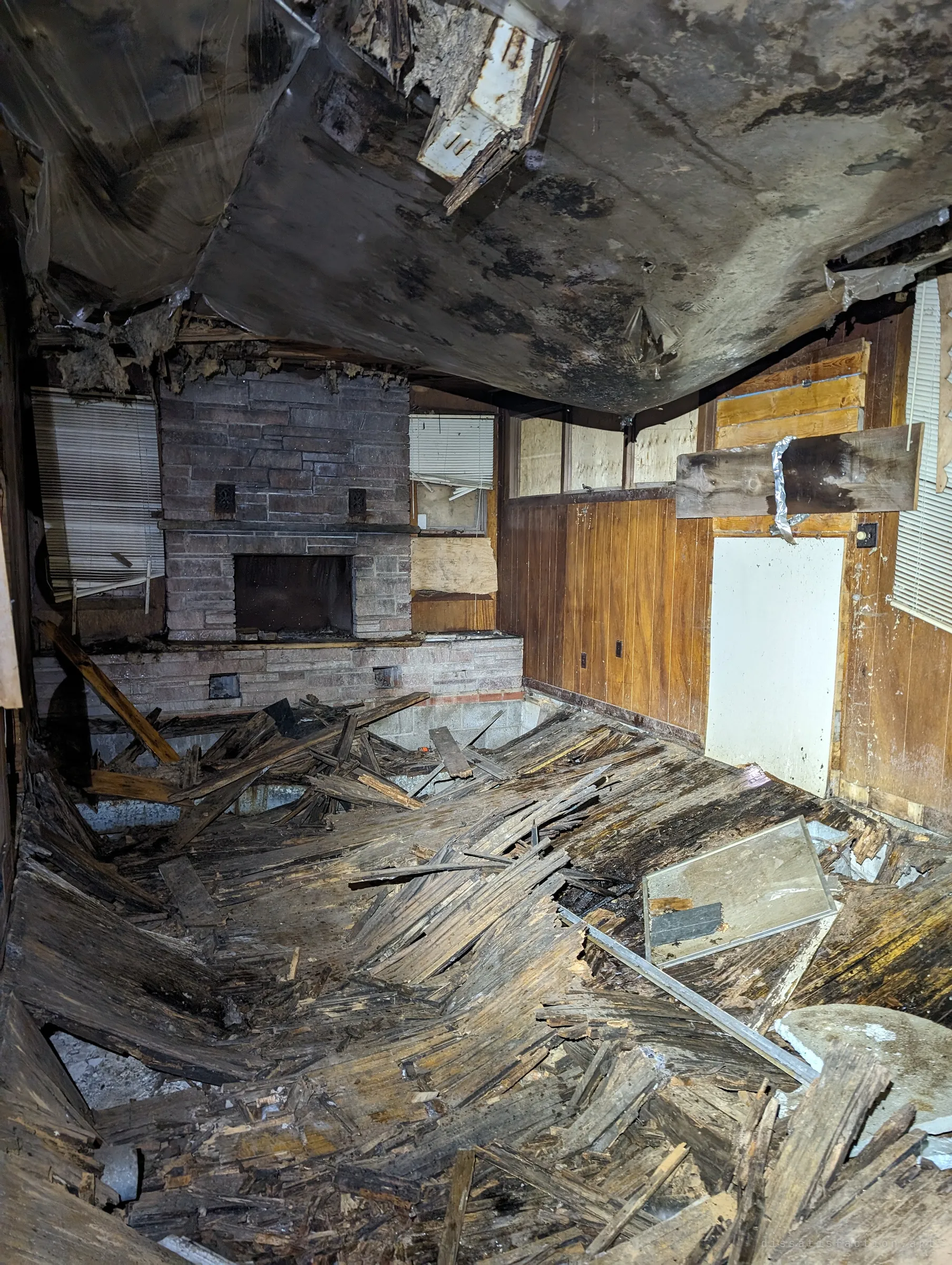 A living room with a stone fireplace where the wooden floor has collapsed into the crawlspace below and the ceiling sags overhead. The rotted boards buckle toward a hole in the middle of the room.