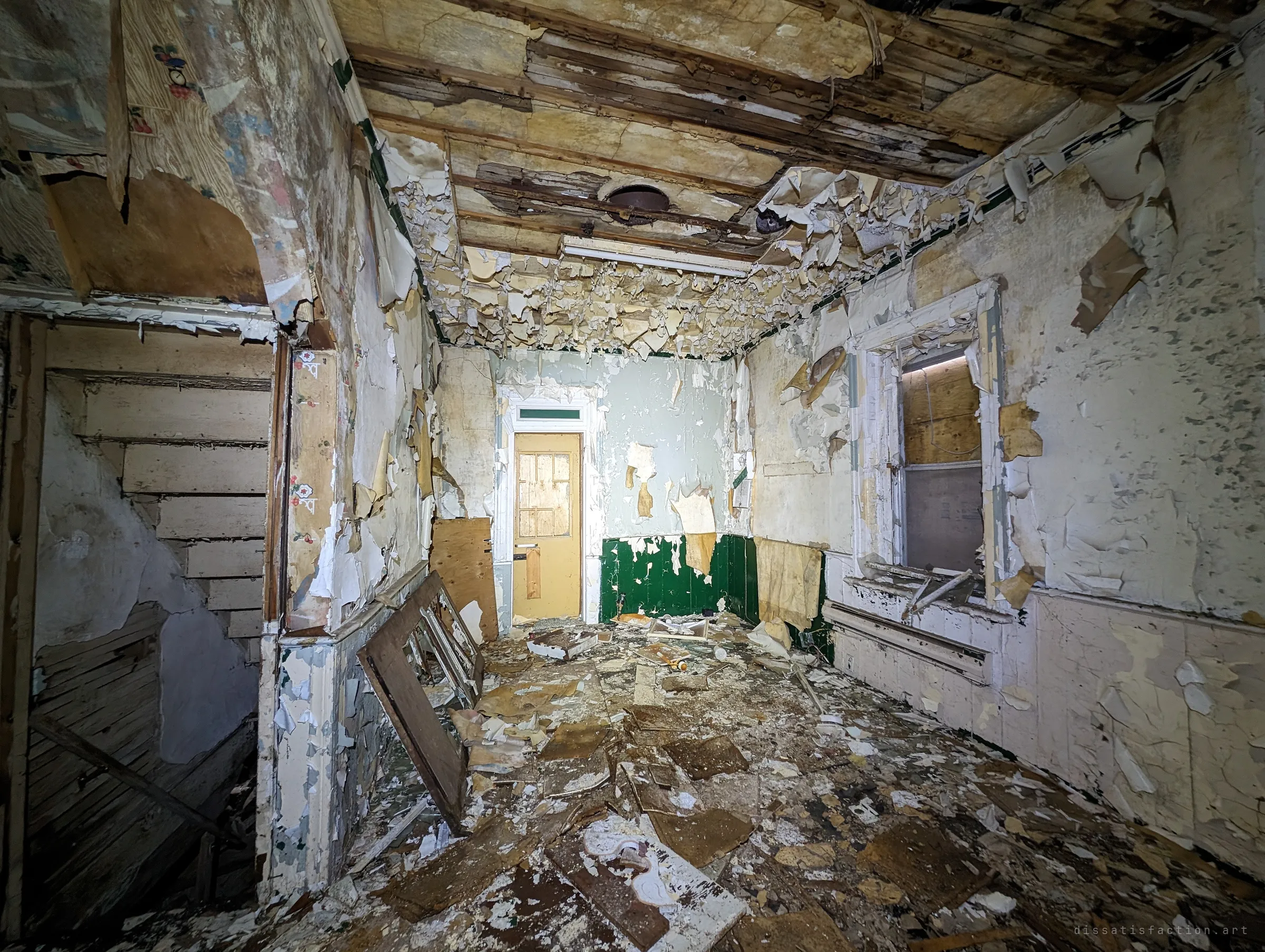 A room where paint and wallpaper hang in curls from every wall and the ceiling, with green wainscoting and a boarded glass-panelled door at the far end. An open stairwell sits to the left.