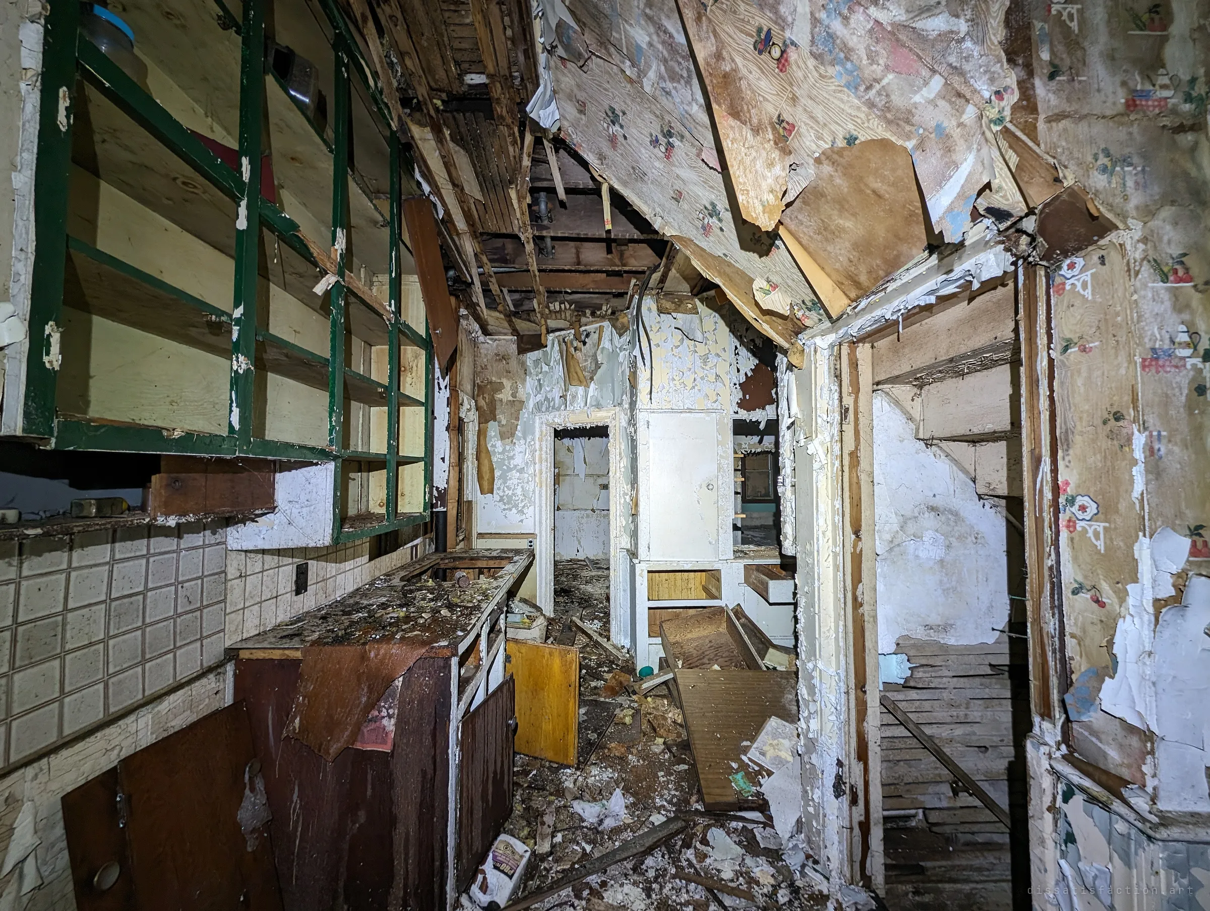 A narrow kitchen with green-framed open shelving, tiled backsplash and gutted cabinets under a partly collapsed ceiling lined with floral wallpaper. The corridor beyond leads past the stairs into more rooms.