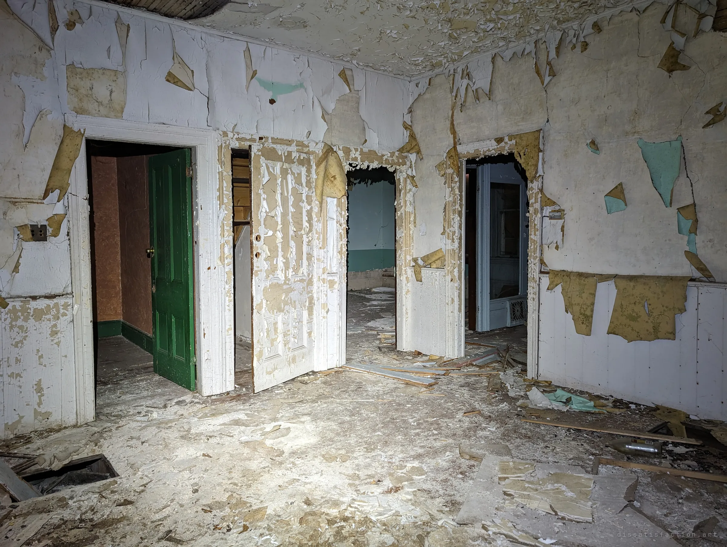 A hall with four doorways, one green door and others white, surrounded by walls shedding layers of paint and paper. The flashlight picks out the debris-strewn floor and a hole cut into the boards.