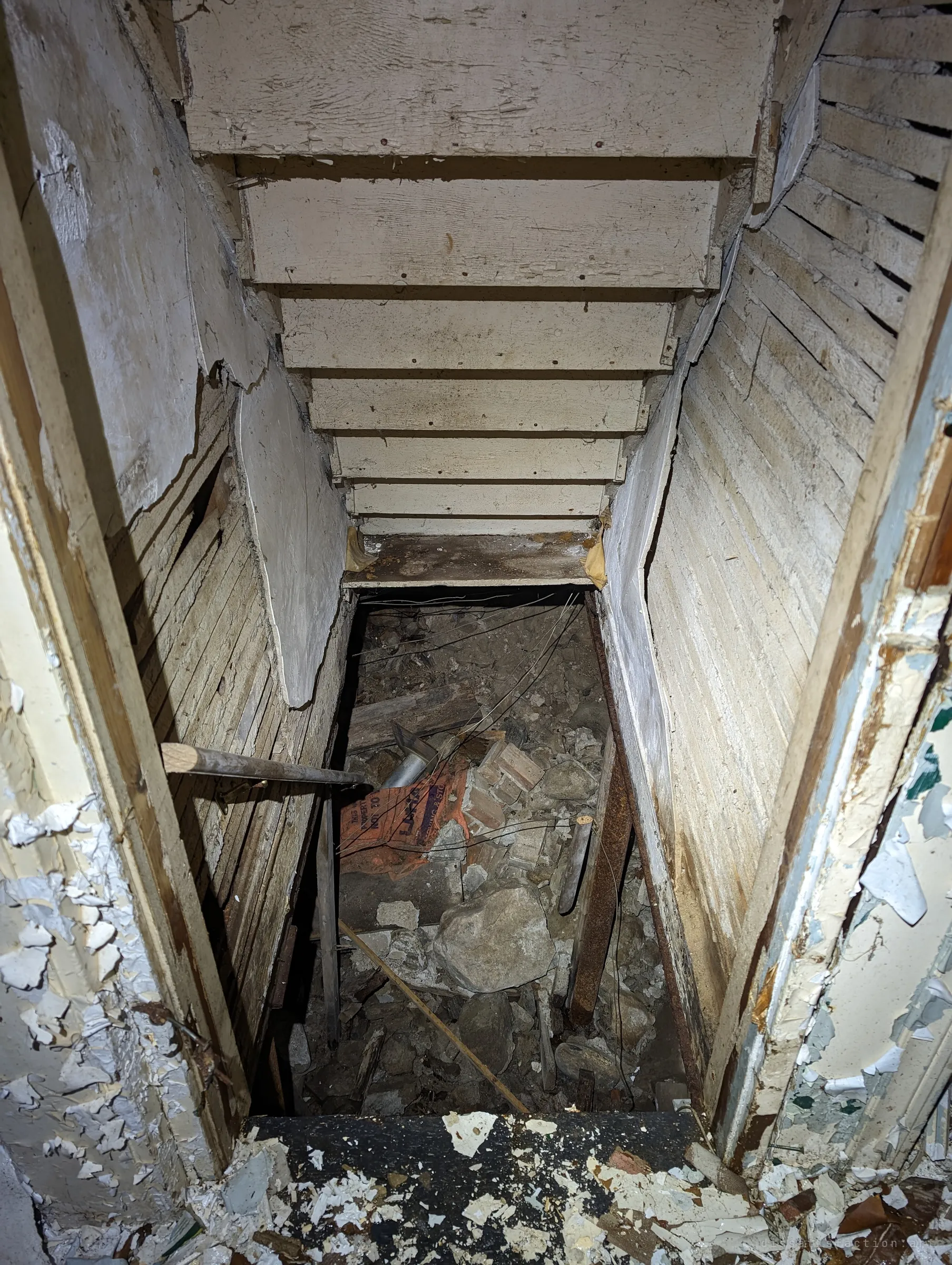 Looking down a narrow cellar stair whose lower steps have gone, leaving a drop into rubble, rocks and broken brick. The cramped white-painted walls make the missing steps feel even more abrupt.