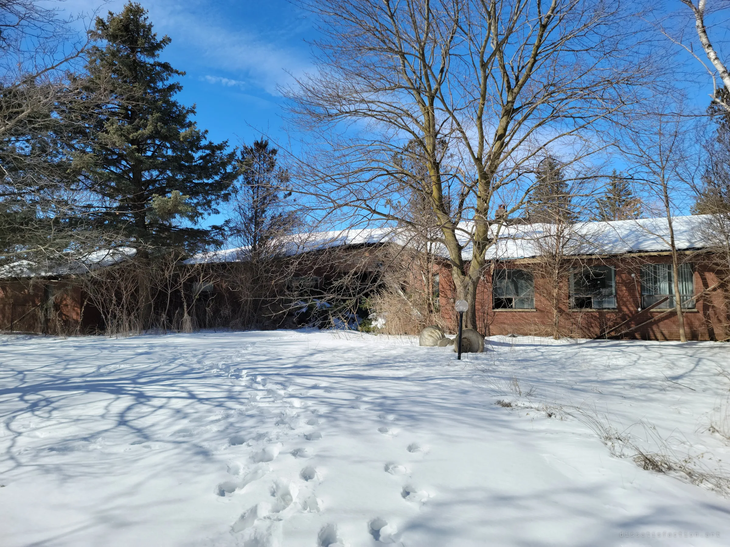 The long, low brick house sits half-swallowed by bare trees behind an untouched snowfield, a lone garden lamp and a line of footprints leading in. It sets the scene: a family home left to winter.