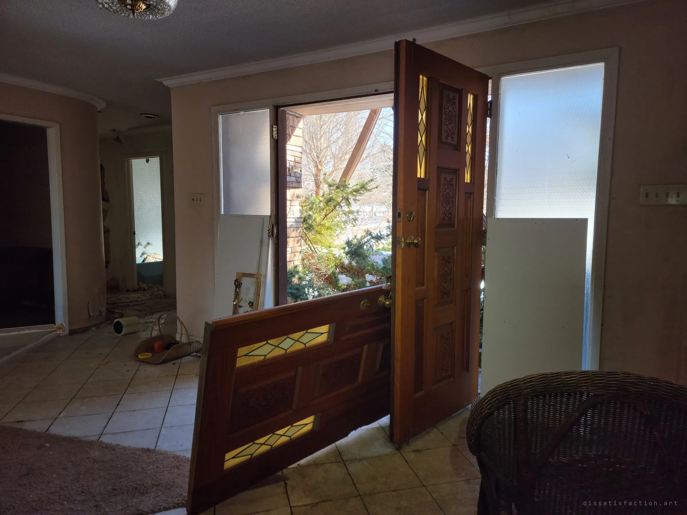 Inside the foyer, a carved wooden door with amber leaded glass hangs off its hinges while the other leaf stands open to the snowy yard. Frosted sidelights and a wicker chair frame the broken entry.