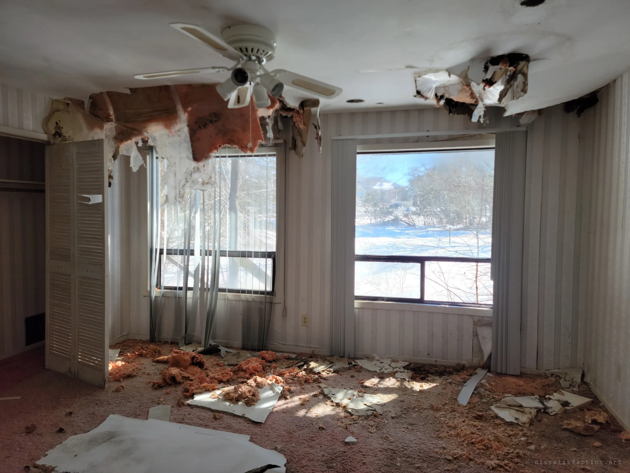 A bedroom with striped wallpaper and a ceiling fan still hanging beneath a ruptured ceiling, insulation dripping onto the carpet. Wide windows look out onto a snowy, overgrown yard.