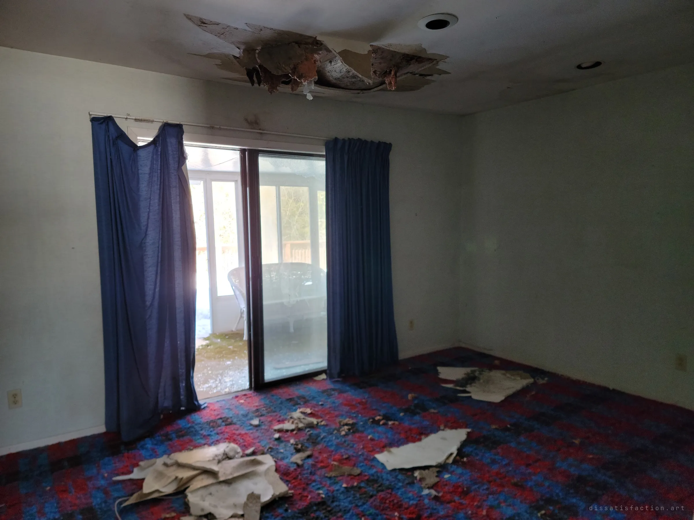 A room with a loud red and blue carpet and navy curtains, a hole blown in the ceiling above. Through the sliding door, a white wicker settee waits on the enclosed porch.