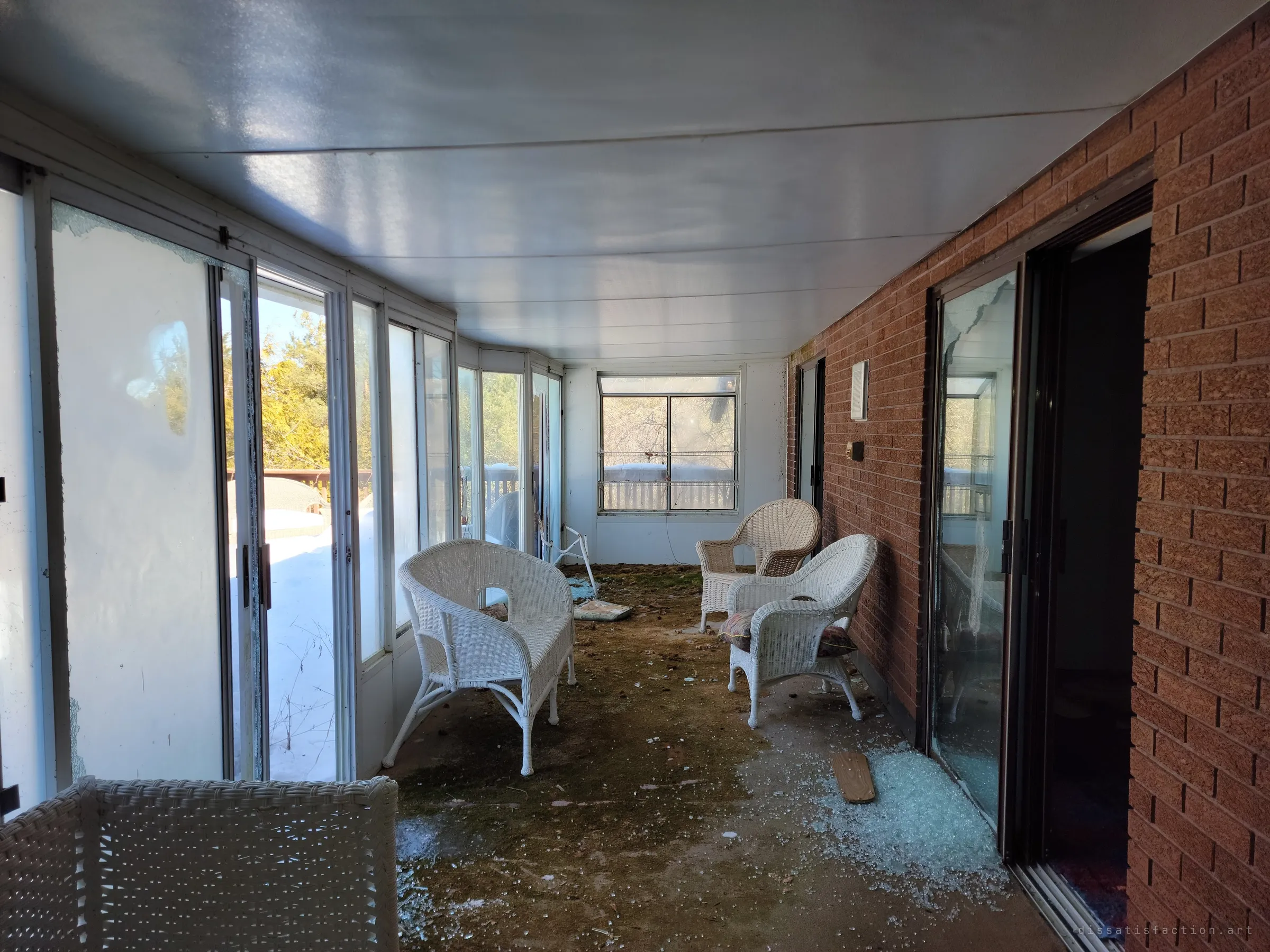 The glassed-in sunroom along the back wall, its white wicker chairs still arranged for company among shattered glass and moss. The brick house wall and a broken patio door line the right side.