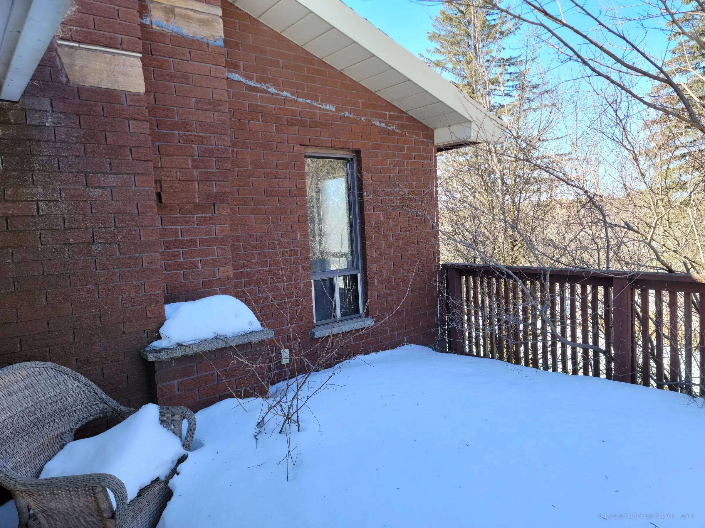 A snow-covered back deck against the red brick gable, a wicker chair buried in drift and trees crowding the railing. The view beyond is only branches and woods.