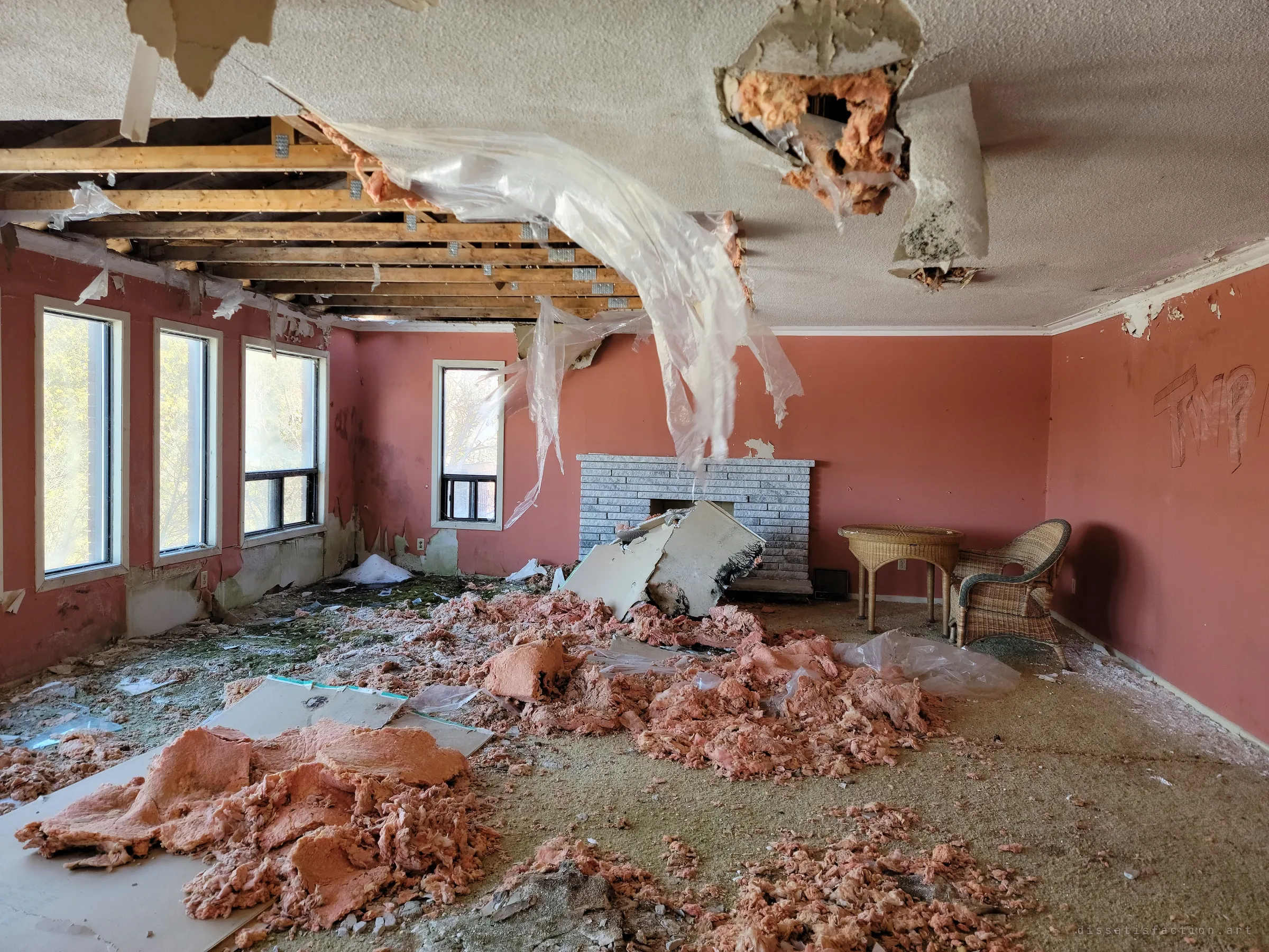 A coral-painted living room with its ceiling stripped to the joists, vapour barrier hanging in sheets over drifts of pink insulation. A marble fireplace, a wicker table and chair, and a graffiti tag on the wall remain.