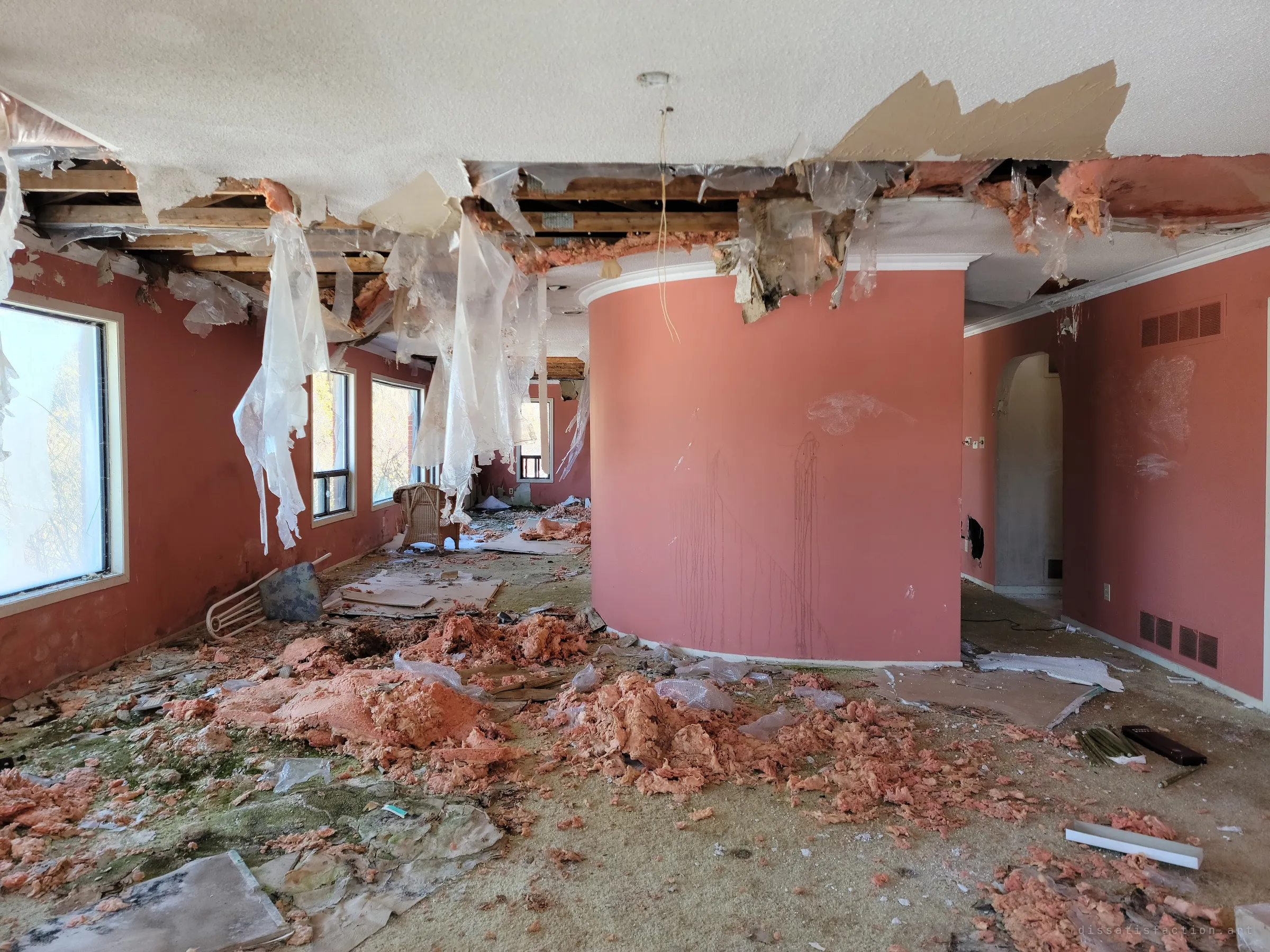 The same coral rooms seen from further back, a curved column wall rising through the wreckage while plastic sheeting hangs from the open ceiling like torn sails. Insulation and moss carpet the floor.