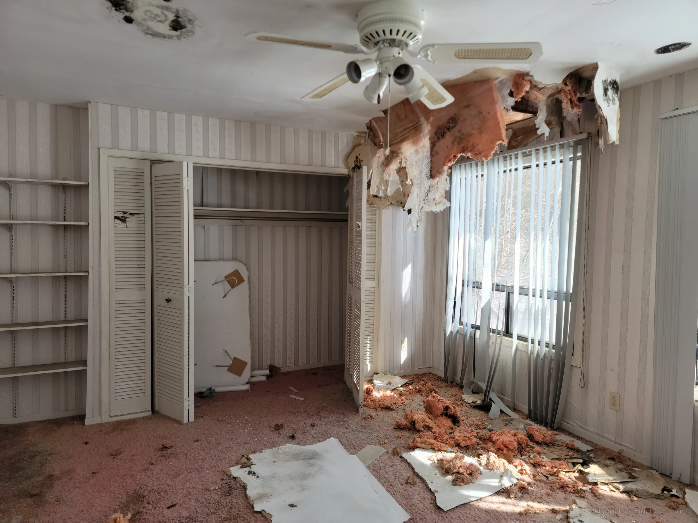 A second striped bedroom with louvered closet doors and empty shelves, its ceiling torn open over the window blinds. A folding table leans in the closet as if put away for later.