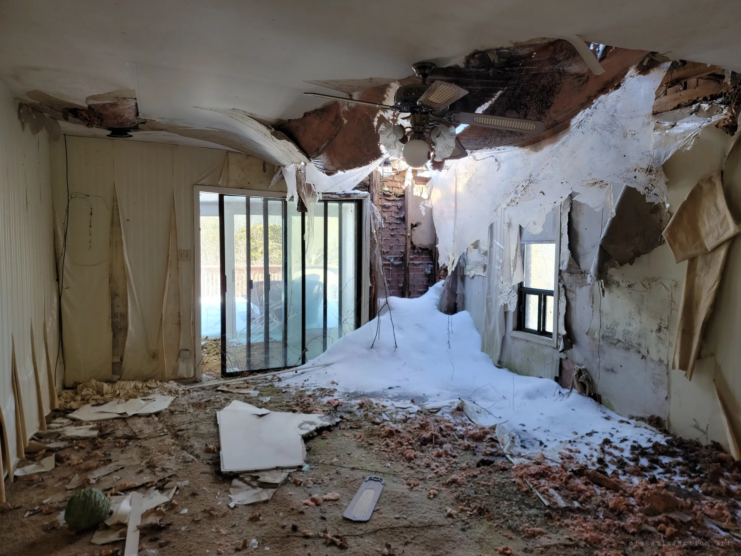 The ceiling has fallen in and snow has drifted right into this room, piling against the wall beneath the exposed brick chimney. A ceiling fan still hangs over the ruin, facing a sliding door to the porch.