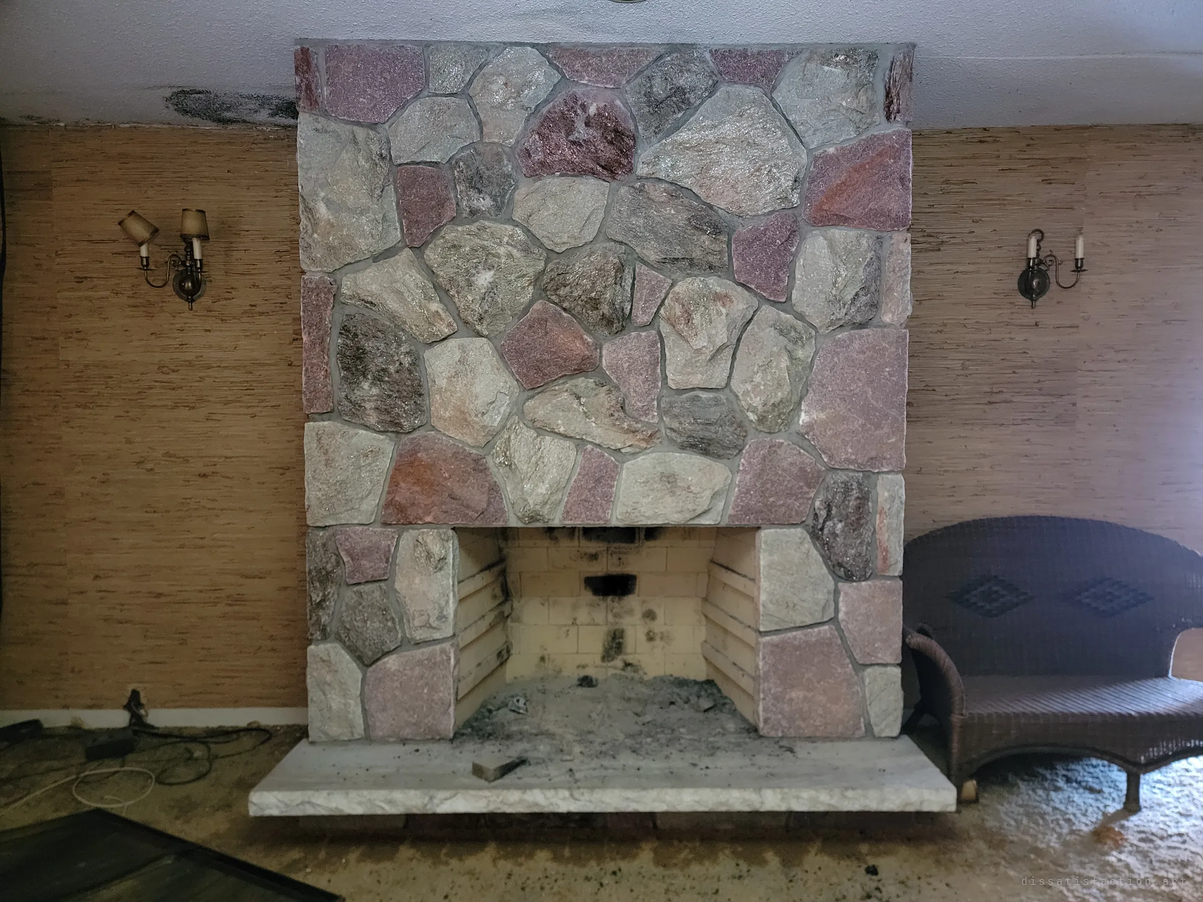 A tall fieldstone fireplace in pink and grey rock fills a grasscloth-papered living room, flanked by brass candle sconces and a wicker loveseat. Soot stains the firebox and mould creeps along the ceiling.