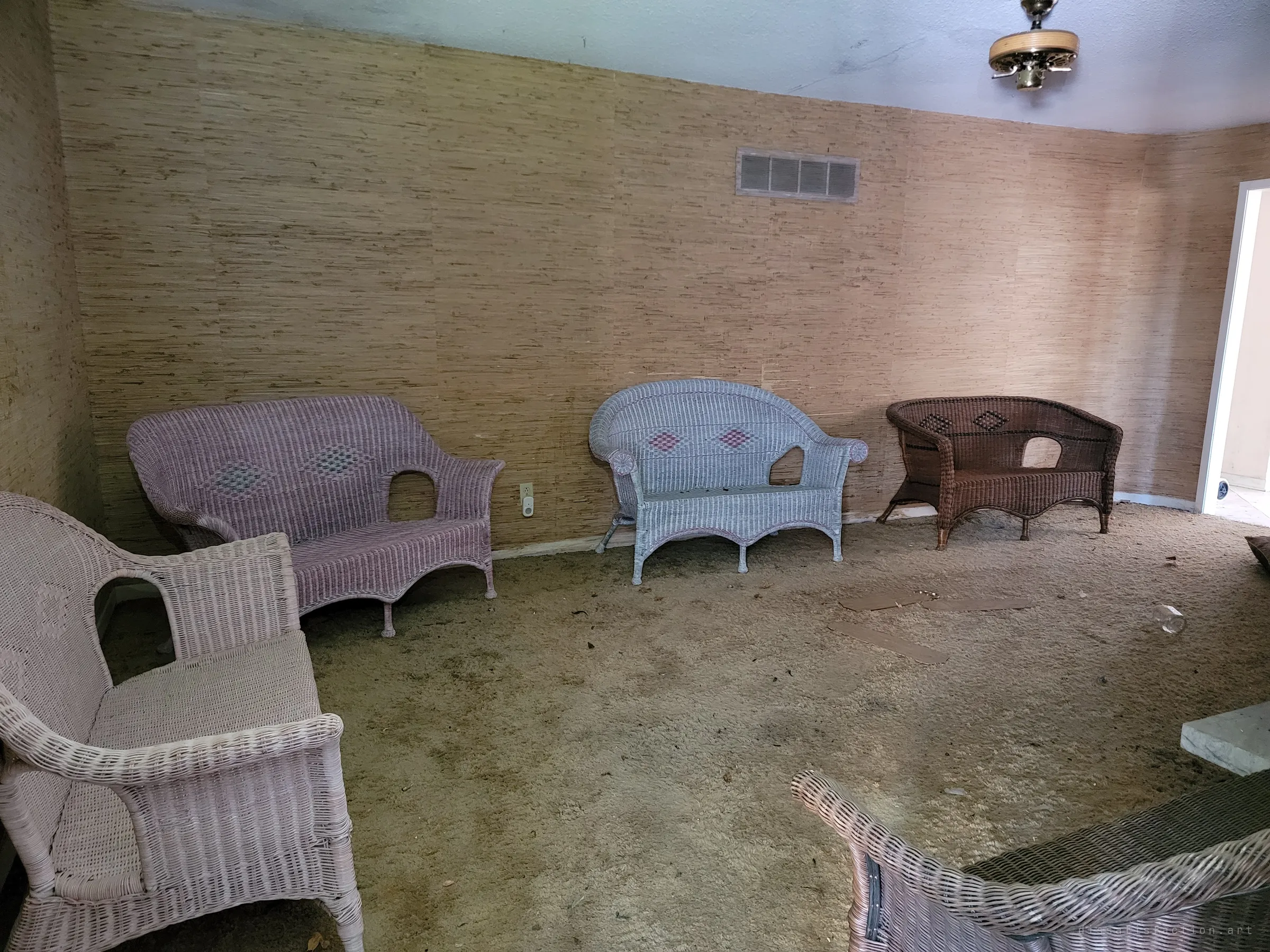 Wicker loveseats in pink, pale blue and brown line the walls of an empty room, as if still waiting for guests. The shag carpet is stained and scattered with debris.