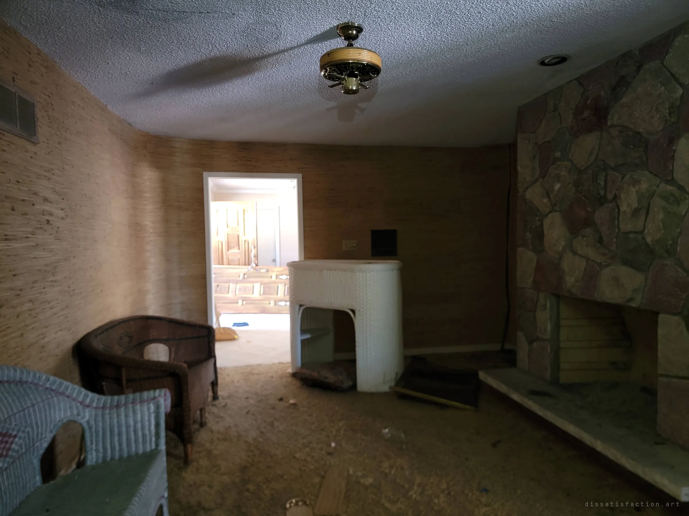 The dim living room seen end to end: a wicker ceiling fan, a white wicker bar and the stone fireplace, with a sunlit doorway opening onto a bedroom beyond.