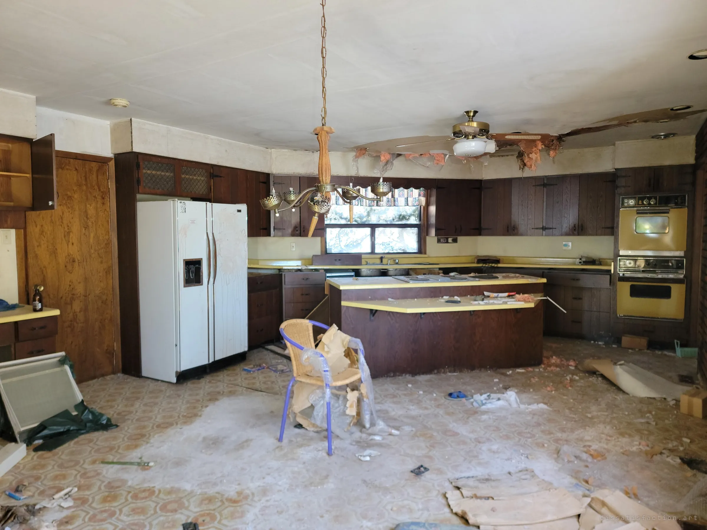 A large dark-wood kitchen with harvest-gold wall ovens, a side-by-side fridge and a central island, its ceiling torn open with insulation hanging down. A lone wicker chair wrapped in plastic sits among the debris.