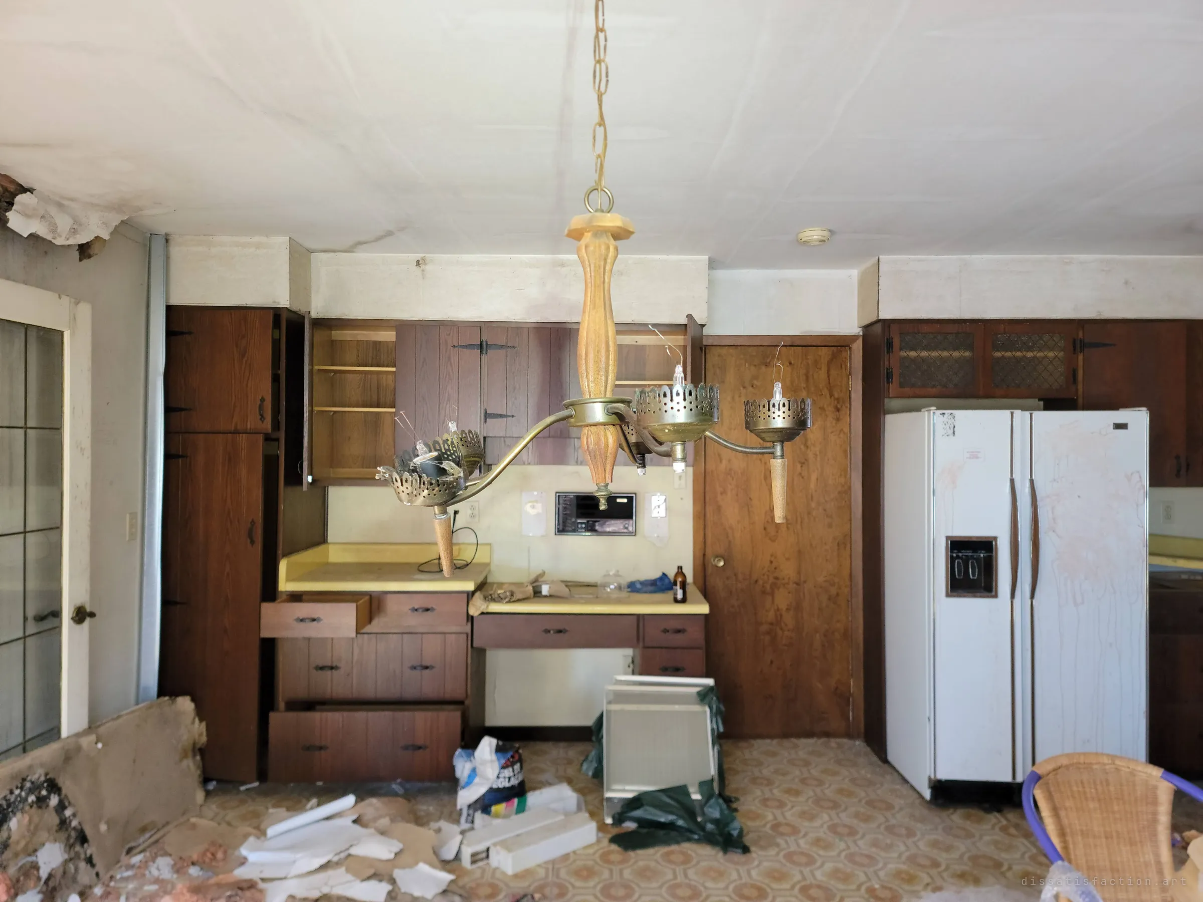 A brass and wood chandelier hangs in front of a kitchen desk nook with an old built-in intercom, pulled-out drawers and a stained white fridge.