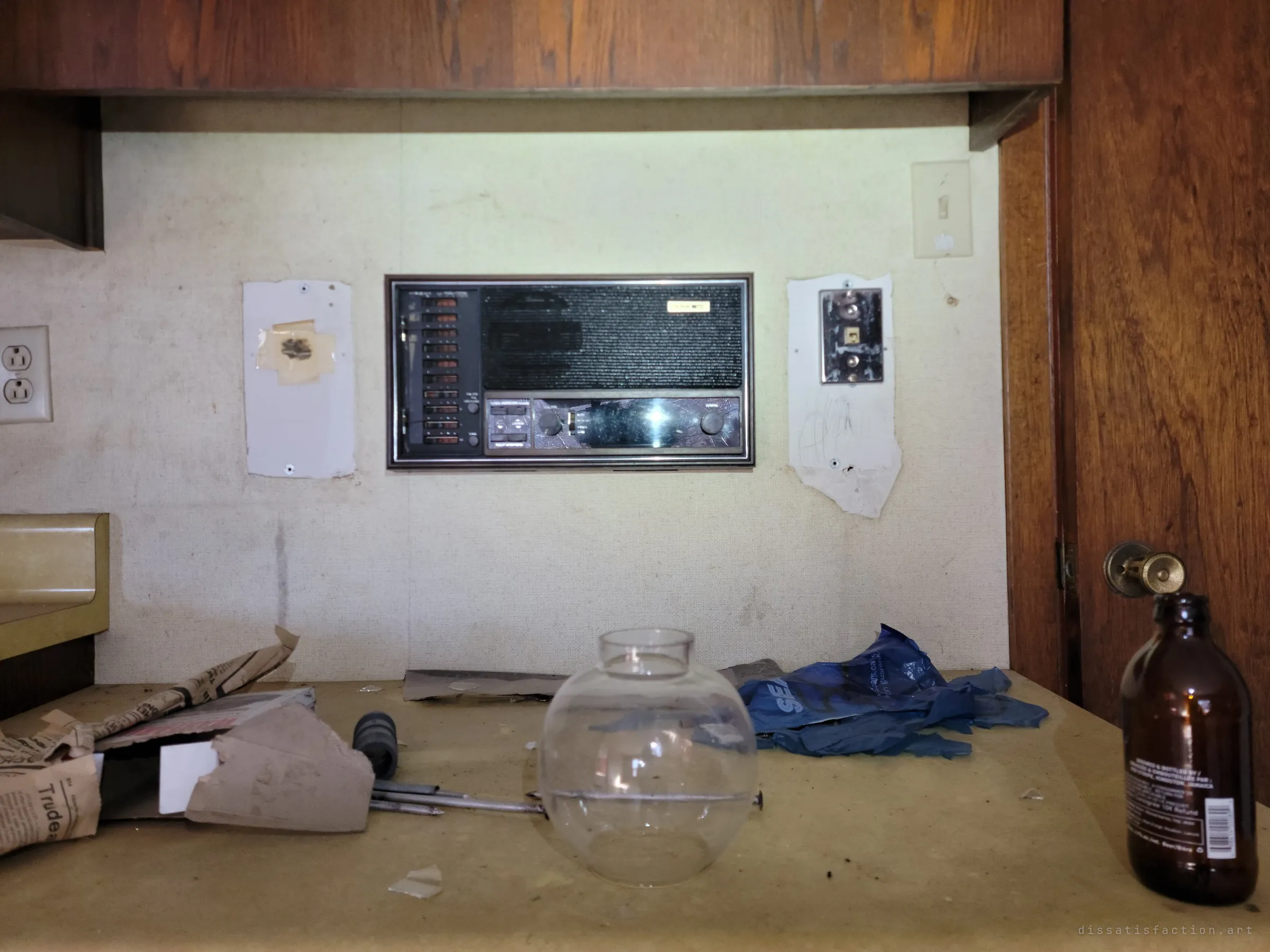 Close on the kitchen desk: a built-in intercom radio panel on the wall above scattered newspaper, a glass lamp globe and an empty brown bottle, frozen from decades ago.